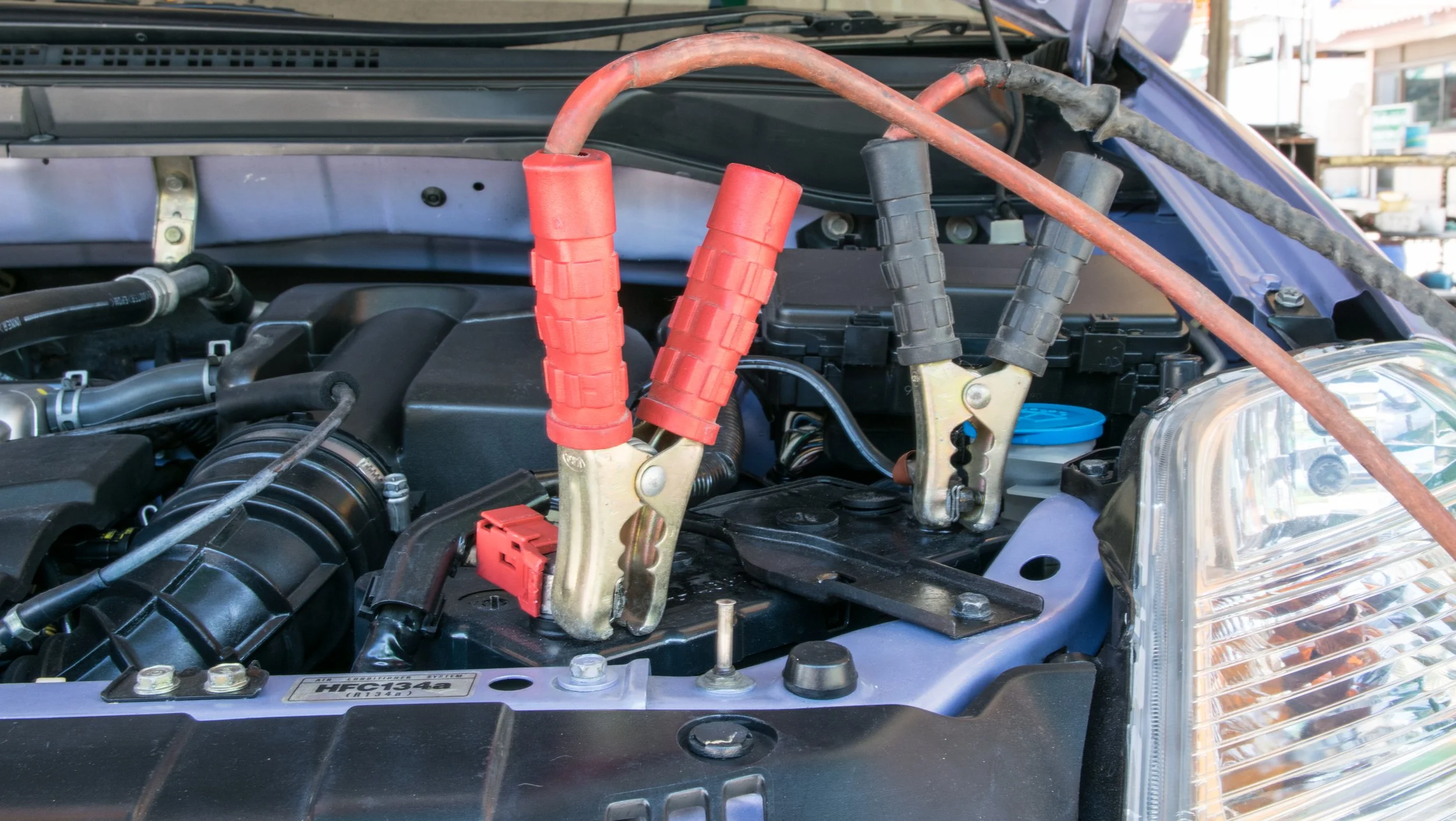 Car hood open with jumper cables connected to the battery, red and black cables attached to the battery terminals.