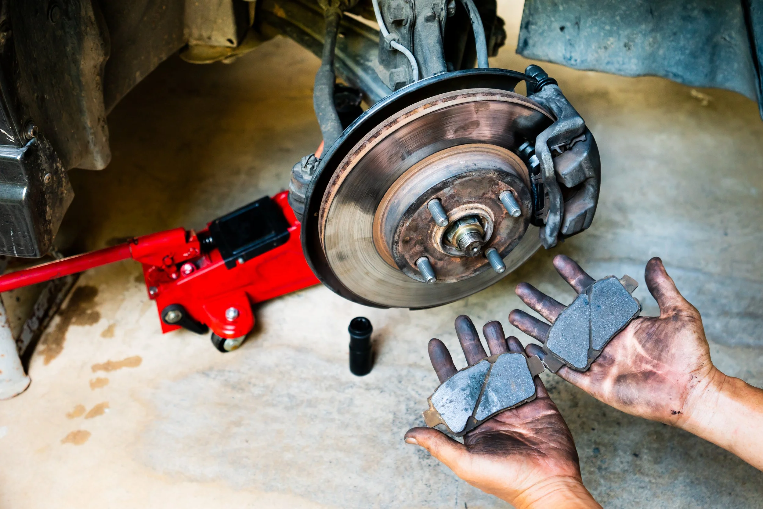 Mechanic holding brake pads near a car's brake rotor which is lifted with a red hydraulic jack, workshop setting.