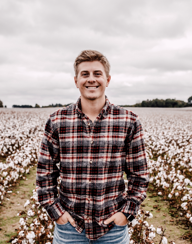 A young man standing in a cotton field during daytime, smiling, wearing a red and black plaid shirt and blue jeans.