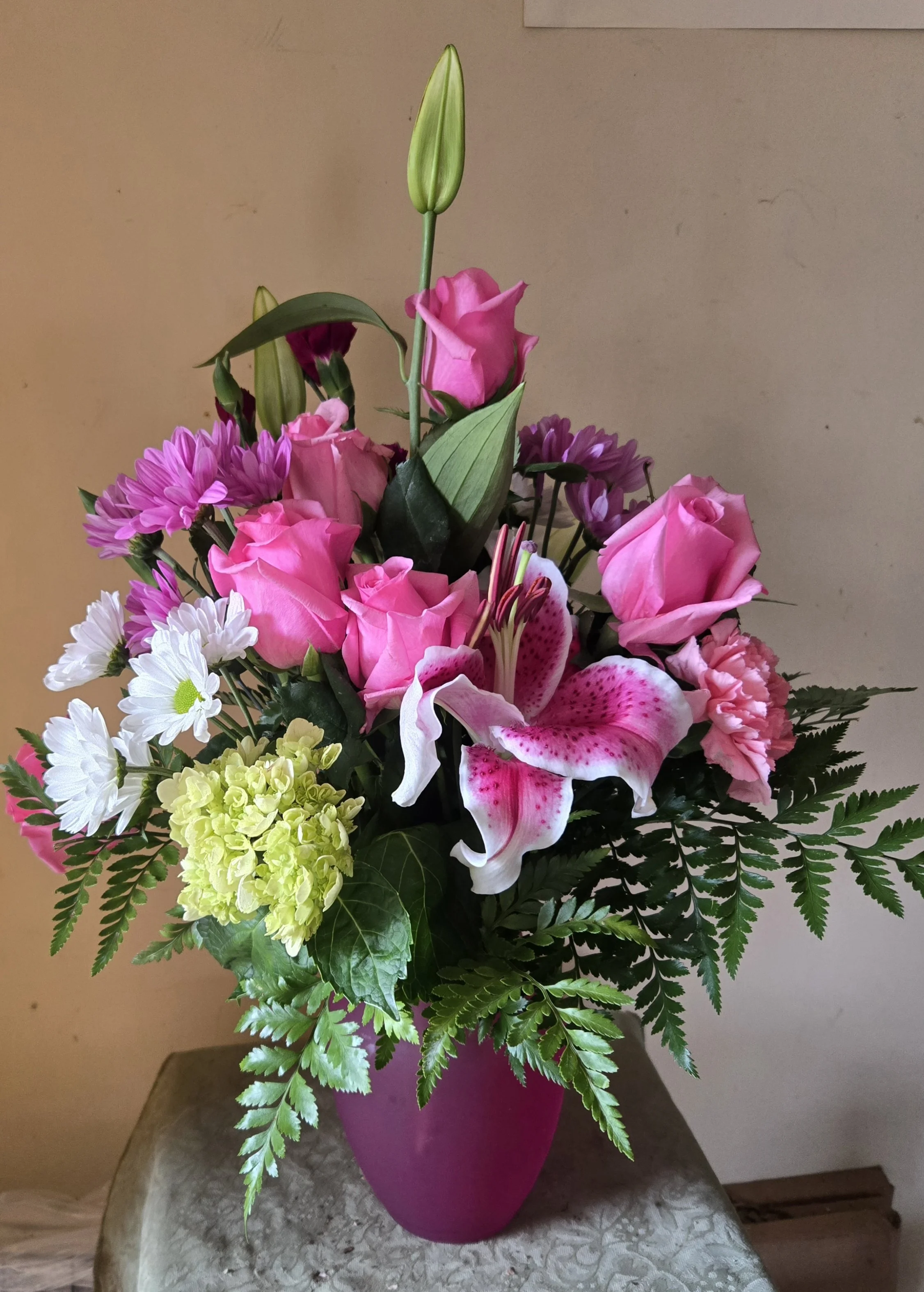 A colorful flower bouquet in a magenta vase with various pink, purple, white, and green flowers and greenery, placed on a textured table against a beige wall.