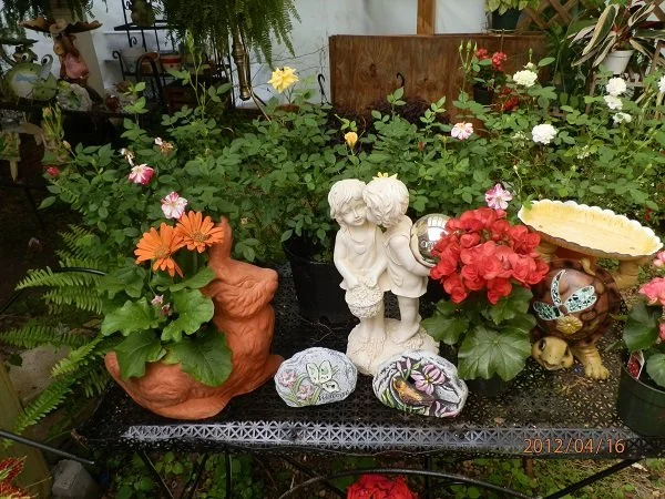 Garden table with decorative garden ornaments, including a brick-like planter with pink and orange flowers, a white statue of children, painted rocks, and various potted plants and flowers.