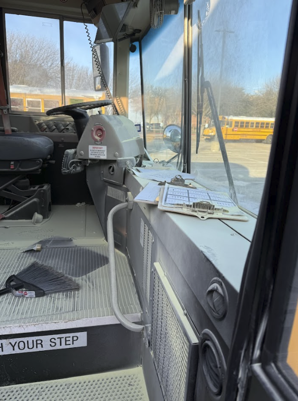 Inside the front of a school bus showing the driver's seat, steering wheel, and a dashboard with clipboards and papers. Outside the large front windshield, a yellow school bus and a parking lot with trees are visible.