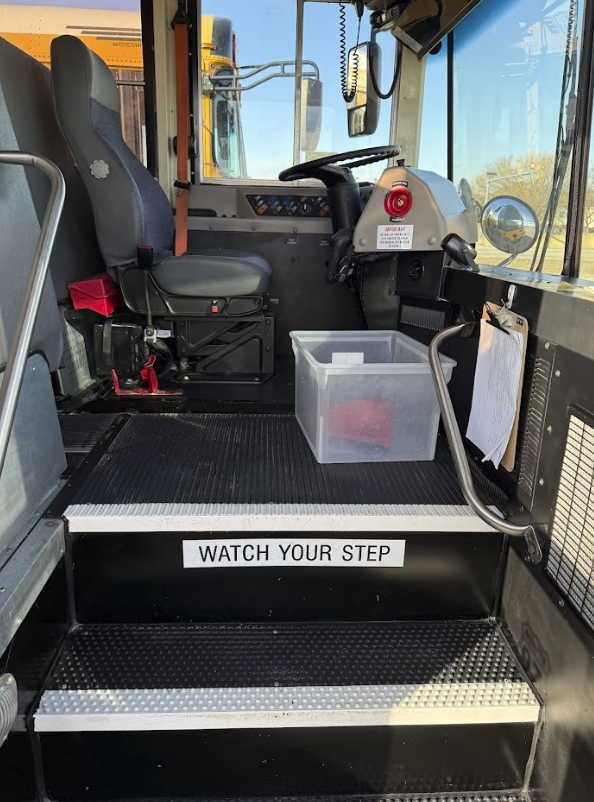 Inside the driver's cabin of a school bus, showing the driver's seat, steering wheel, and control panel. A plastic bin is placed on the floor near the steering wheel. A warning sign on a step reads 'WATCH YOUR STEP.'
