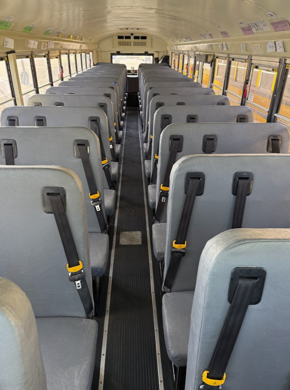 Empty school bus interior with rows of gray seats and seat belts, viewed from the rear looking toward the driver's area.