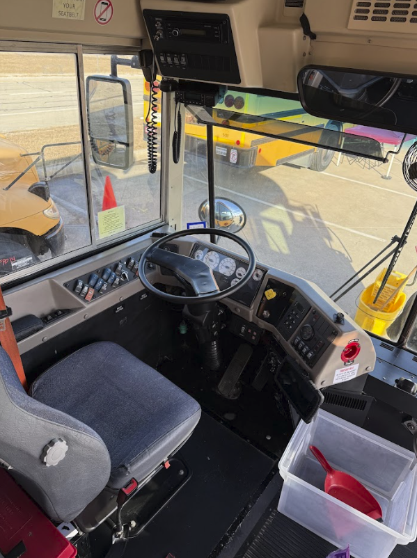 Interior view of a school bus driver's area showing a gray seat, steering wheel, dashboard with controls, and a clear plastic container with a red scoop inside placed on the floor.