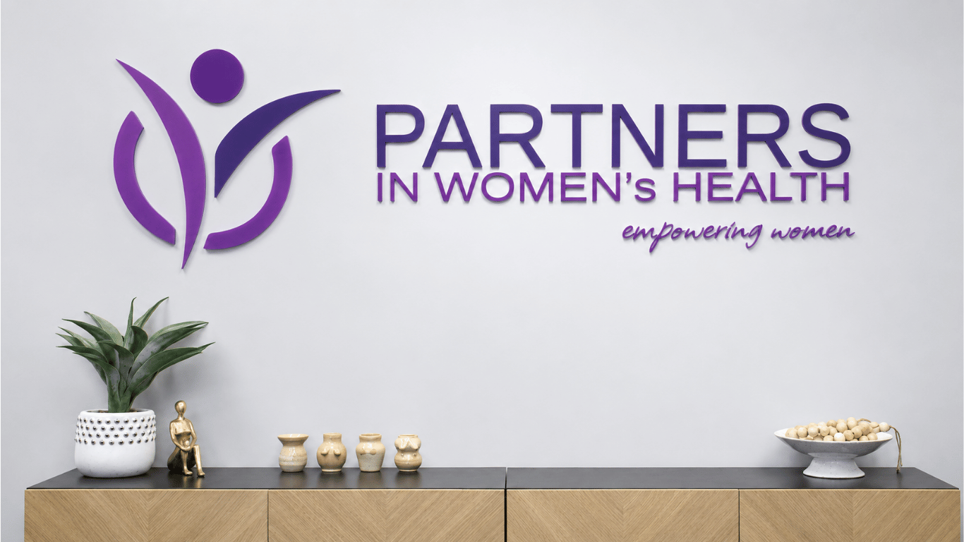 A reception desk displays a potted snake plant, a small figurine, several beige vases, and a bowl of white objects. Behind, a white wall features the purple logo and text for 'Partners in Women's Health' with the tagline 'empowering women'.