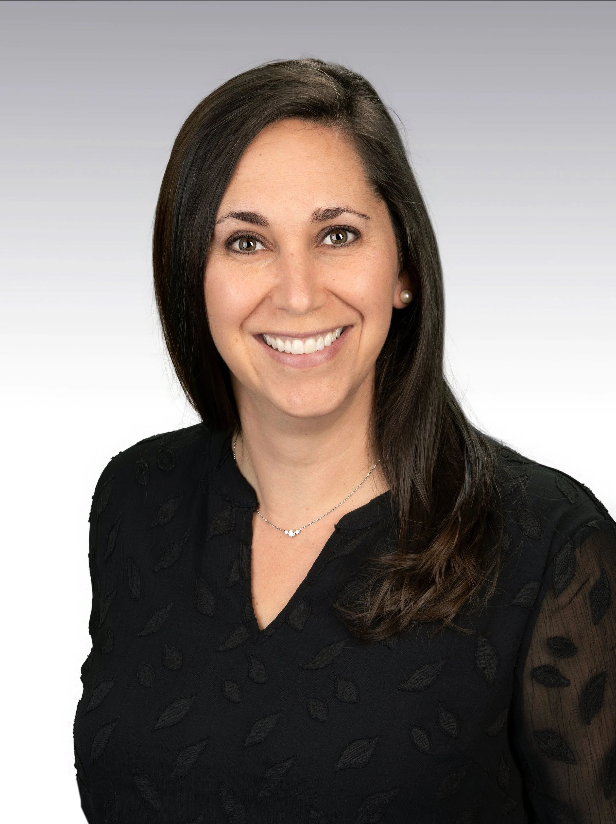 Portrait of a woman with dark brown hair, fair skin, green eyes, and pearl earrings, smiling and wearing a black embroidered top and a delicate necklace, against a plain gray background.