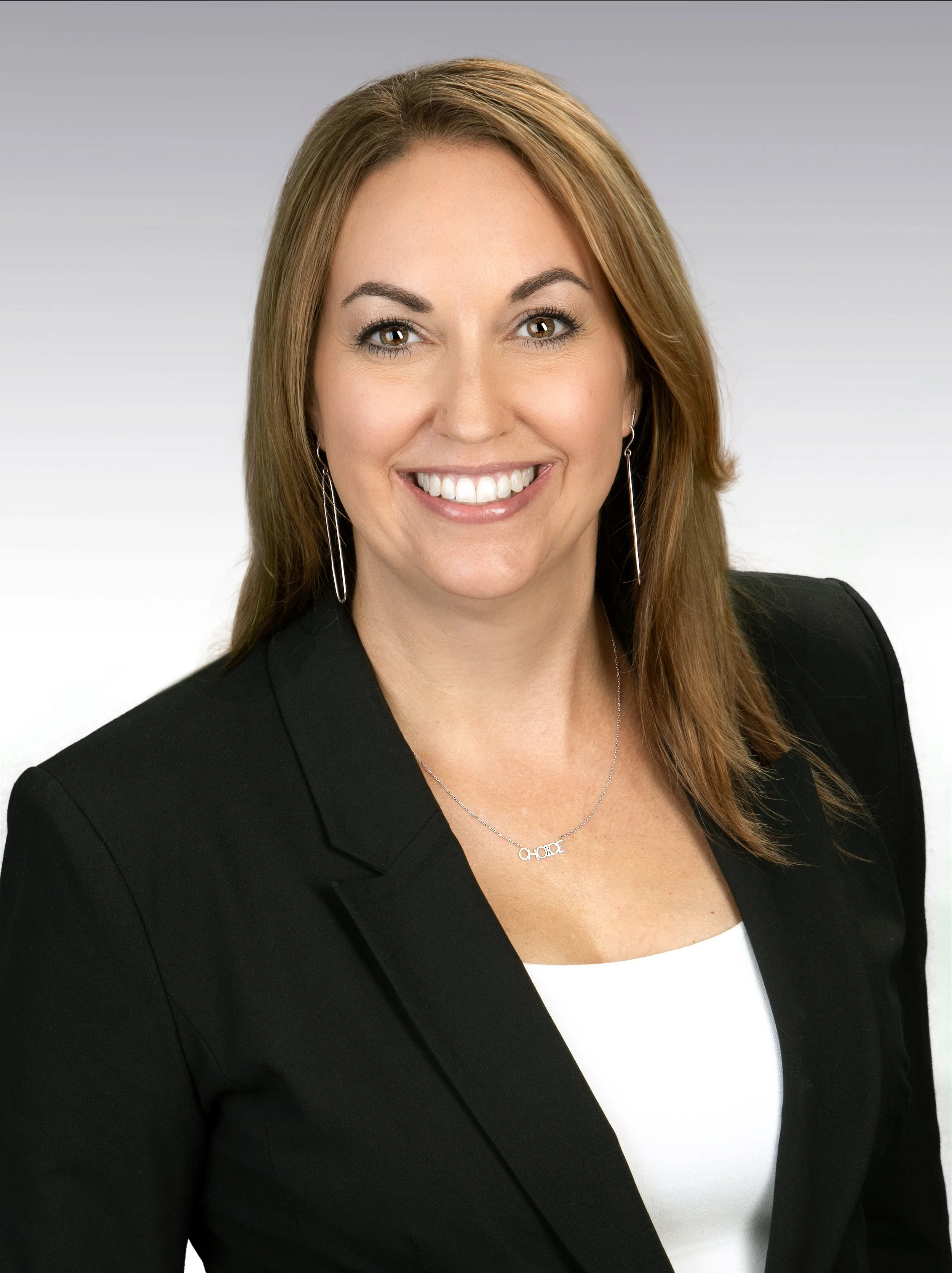 A smiling woman with light brown hair, light makeup, wearing a black blazer, white top, hoop earrings, and a necklace that spells 'CHOOSE', against a gray and white gradient background.