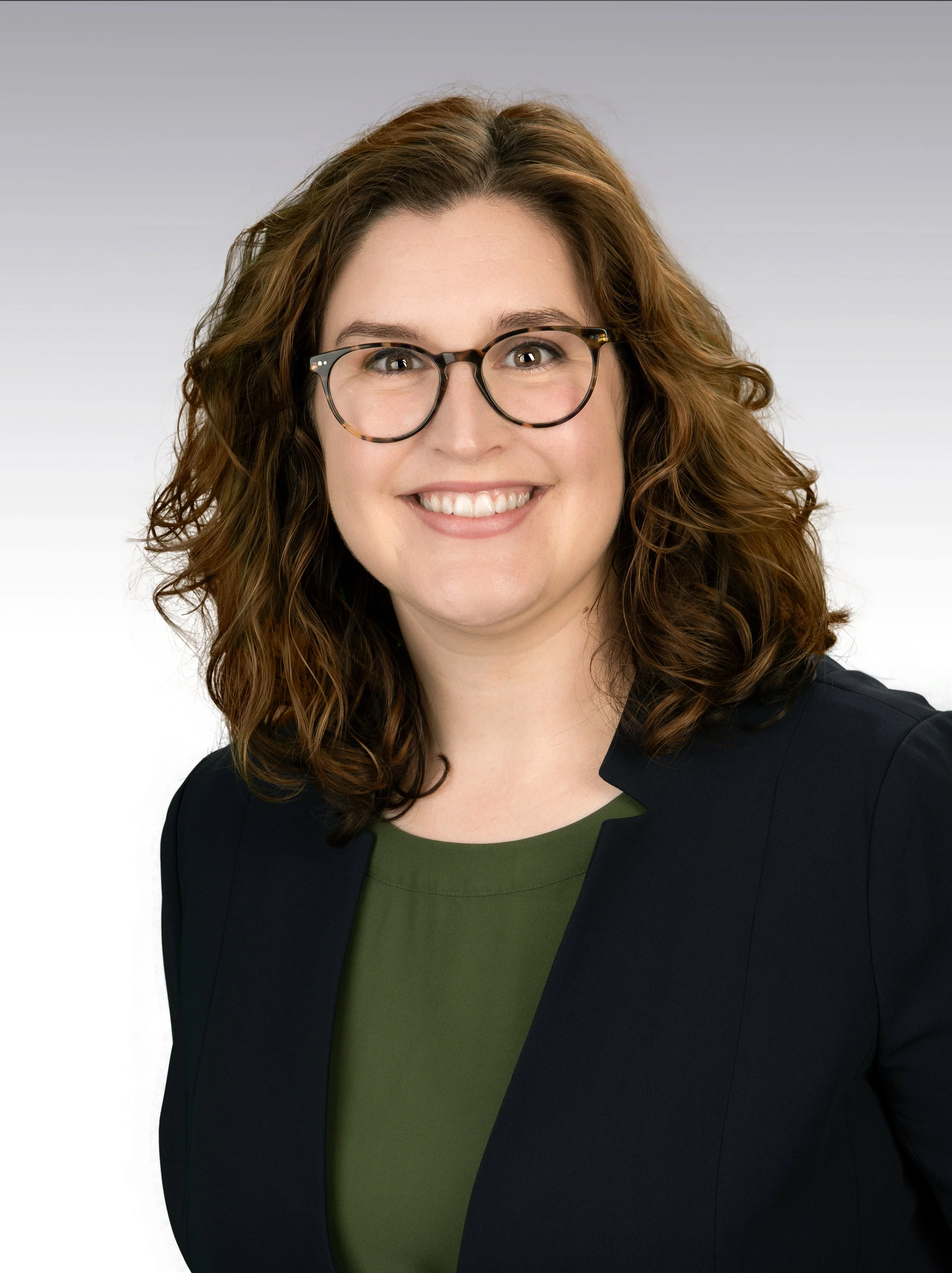 A professional woman with shoulder-length wavy brown hair and glasses, smiling in a studio with a plain, light background.