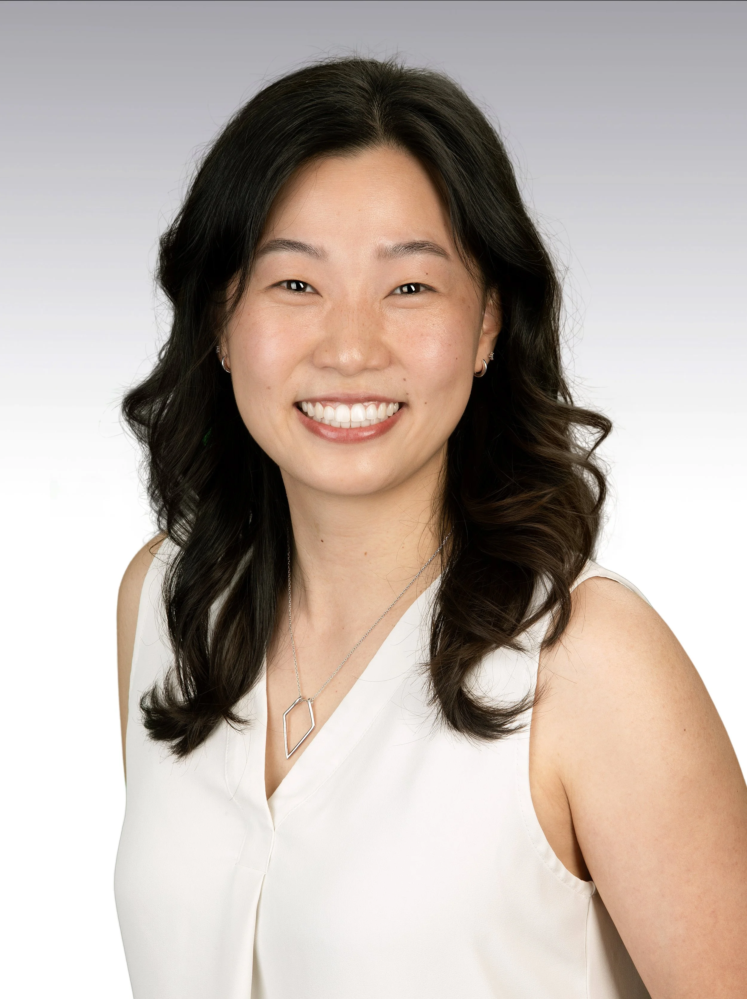 Close-up portrait of a smiling woman with long dark wavy hair, wearing a white sleeveless top, silver jewelry, and light makeup, against a gray gradient background.