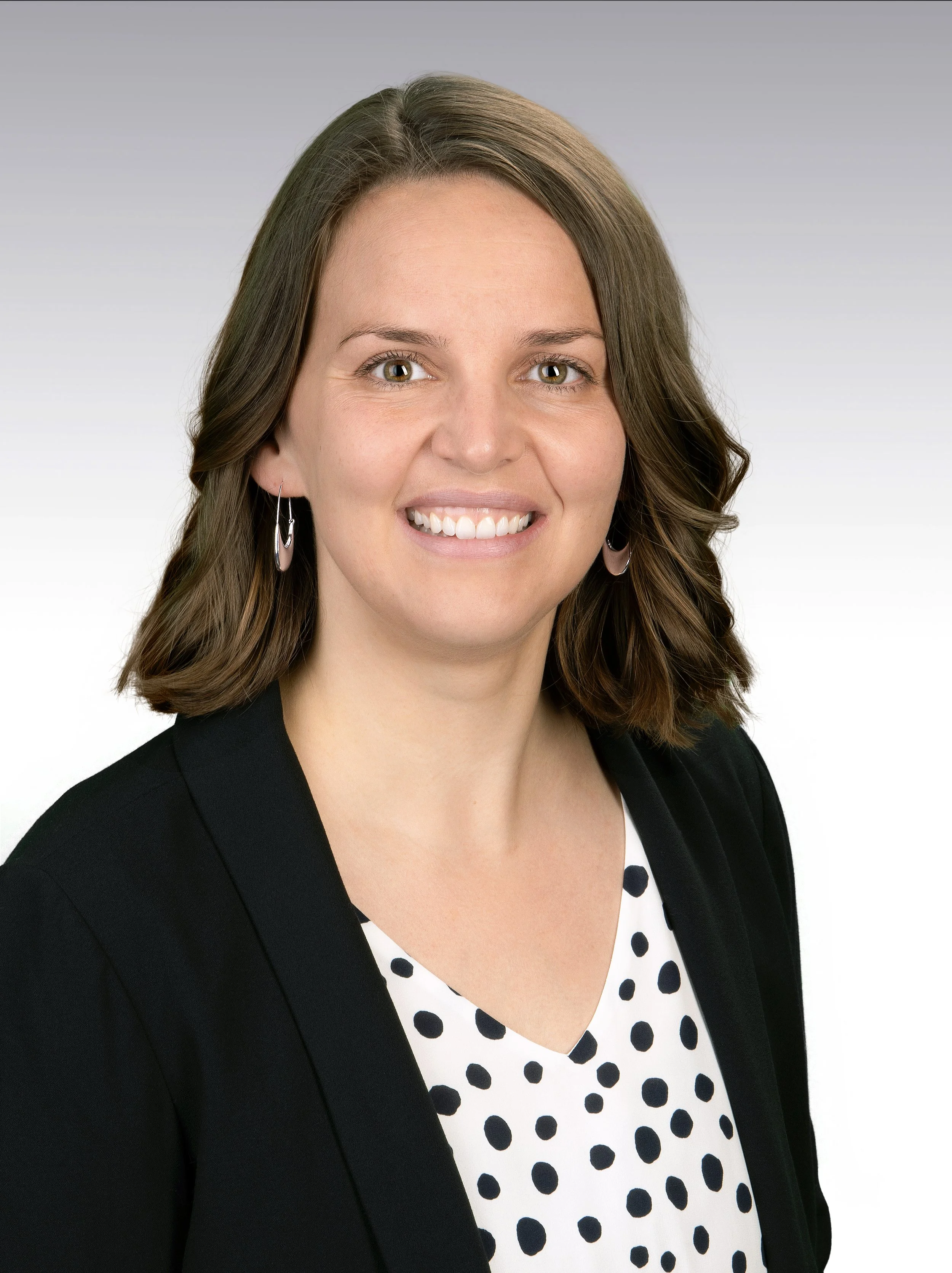 A woman with brown hair styled in loose waves, wearing a black blazer over a white polka dot blouse, smiling with teeth showing, and wearing hoop earrings.
