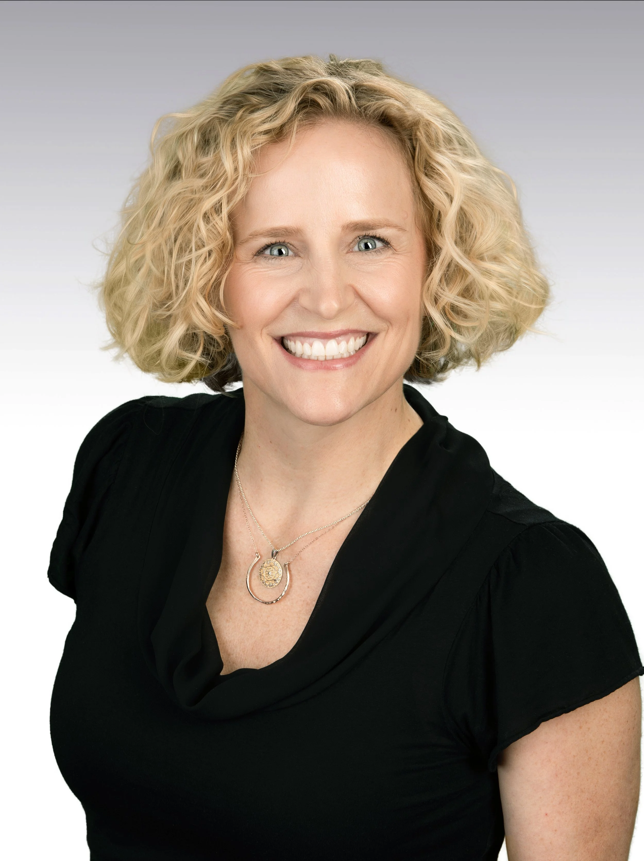 Close-up portrait of a smiling woman with blonde curly hair, wearing a black top and layered necklaces, against a plain light gray background.