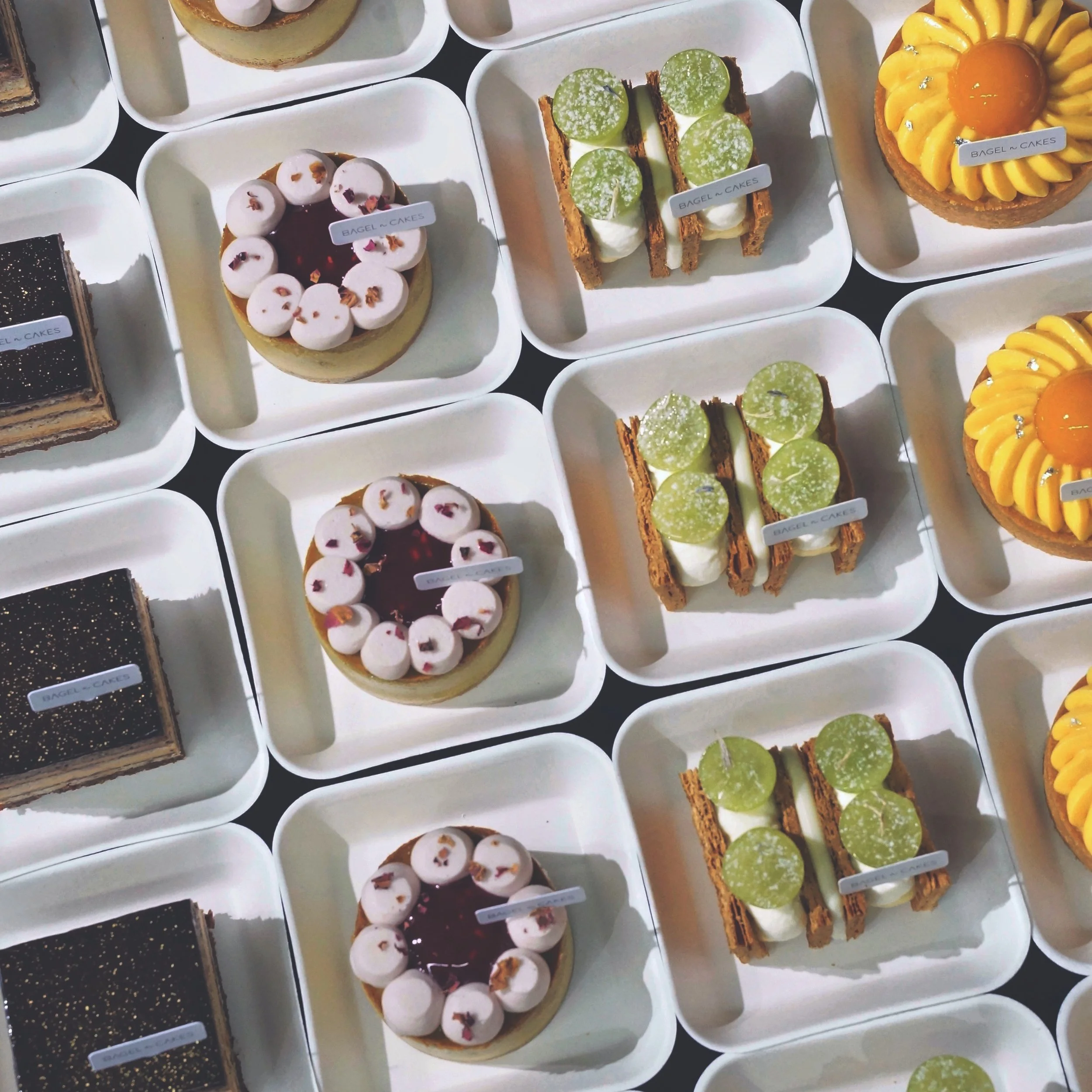 Four types of individual pastries on white trays, including chocolate layered cakes, round raspberry cakes, rectangular mille feuille with green grapes, and round tarts with yellow cream and mango jelly in the center
