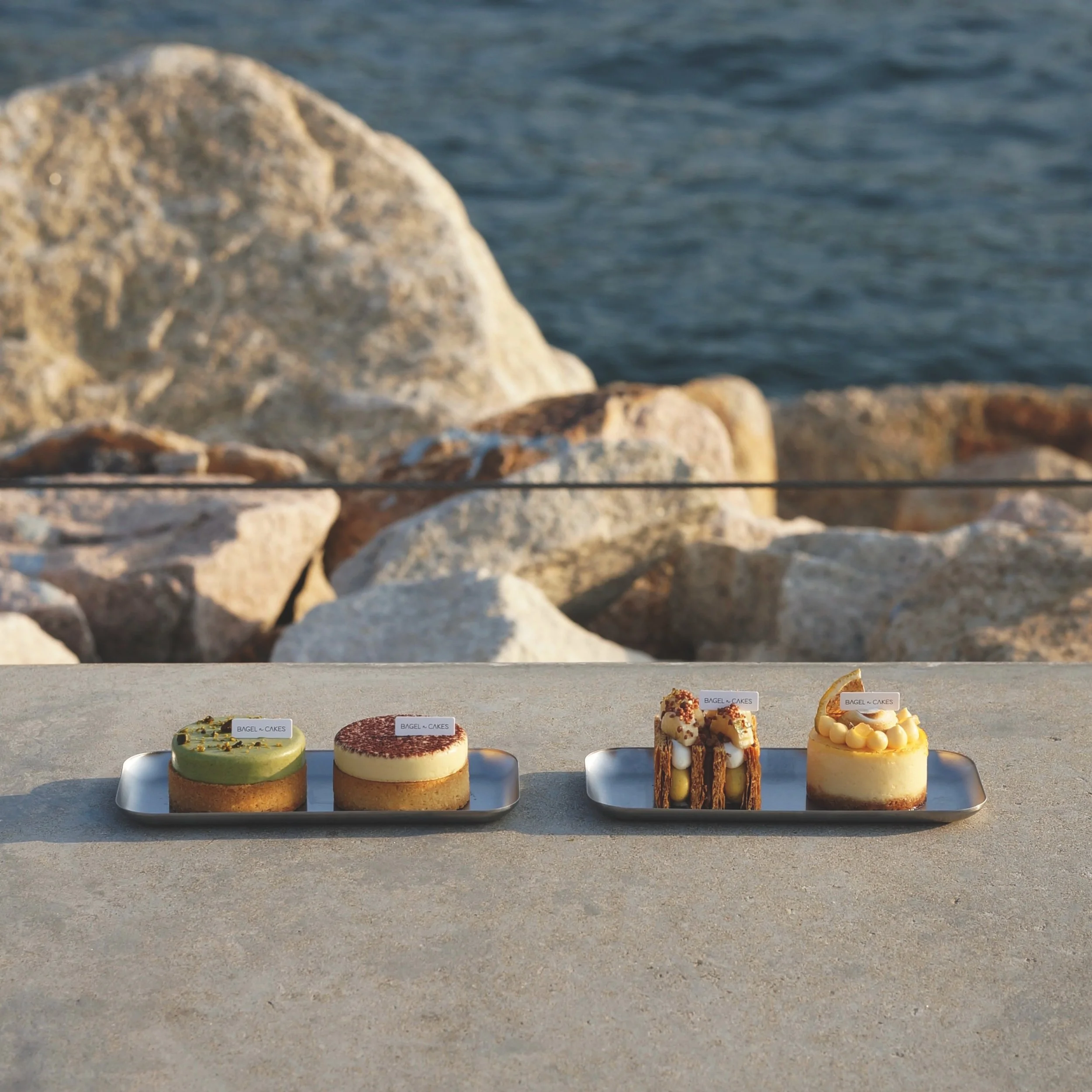 Four small, dessert cakes on metal trays, arranged on a concrete surface in front of the harbor