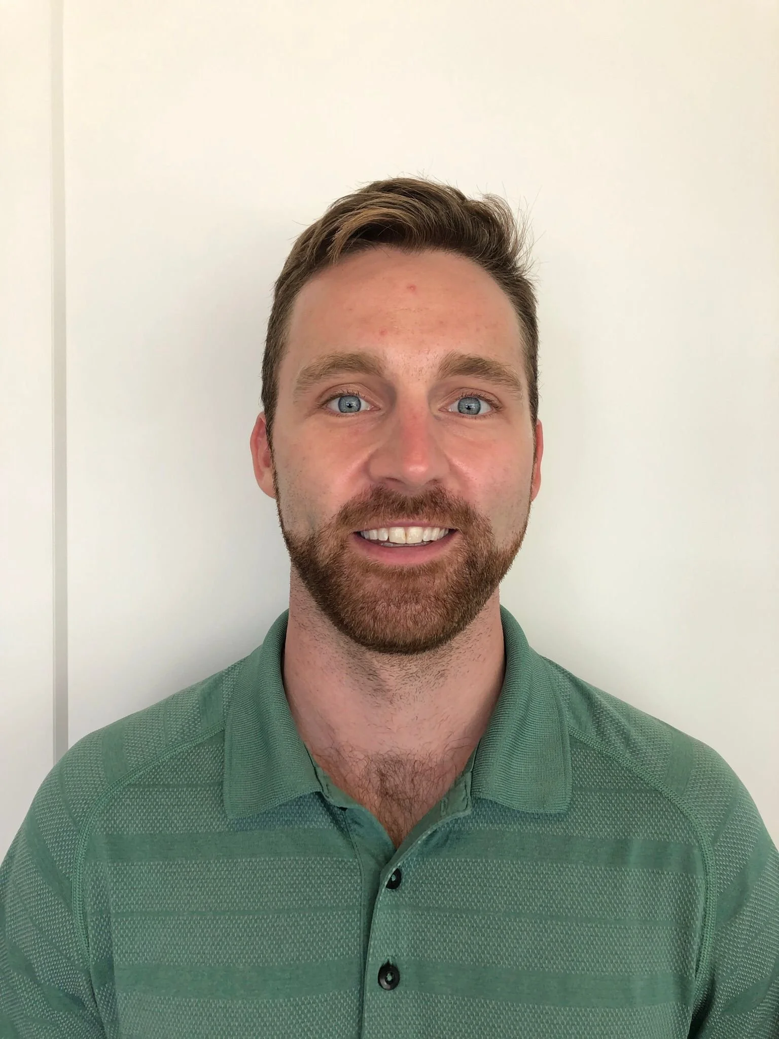 Close-up of a smiling young man with blue eyes, brown hair, and a beard, wearing a green collared shirt, standing against a plain white wall.