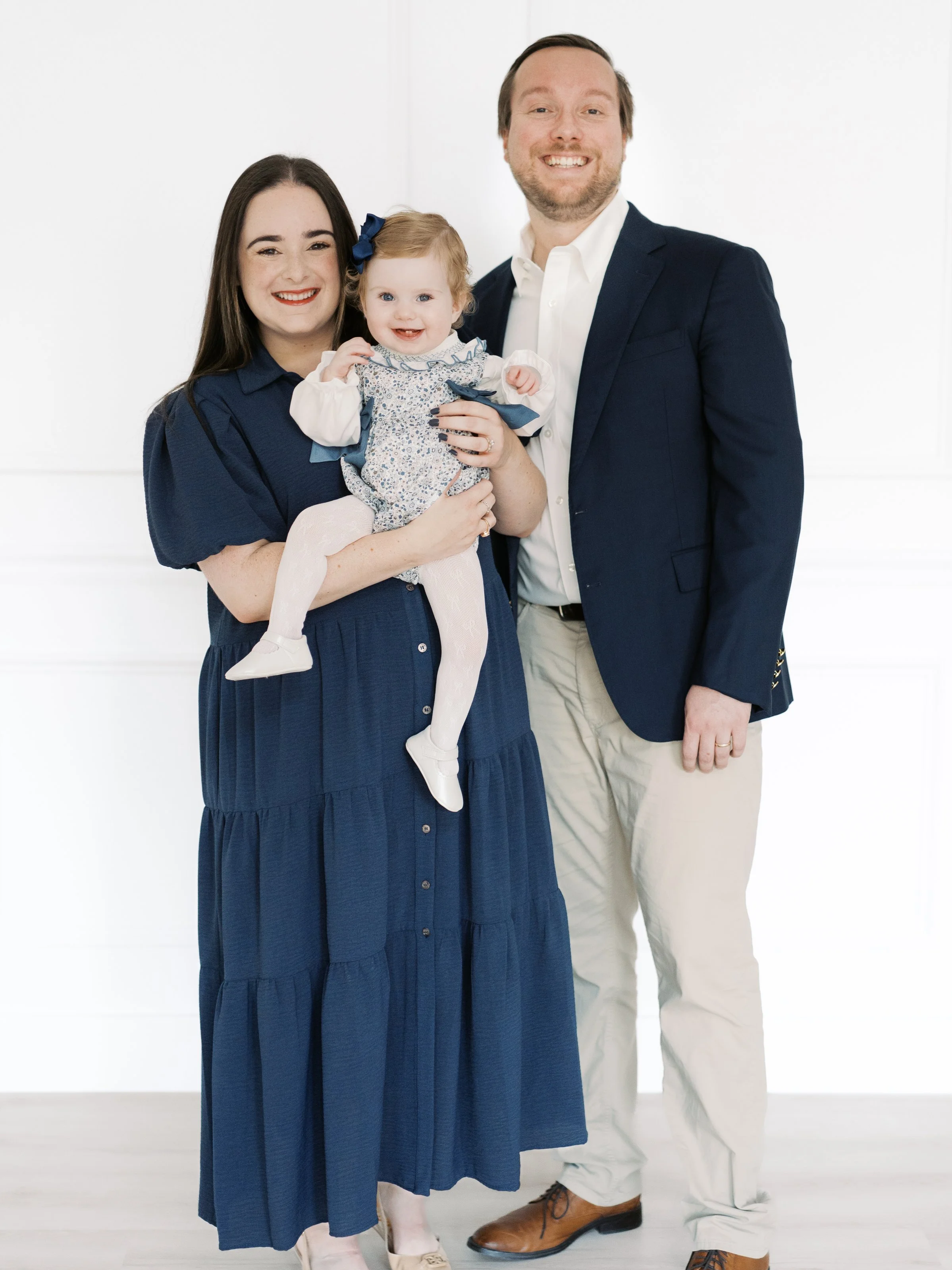 A family of three smiling, with a woman holding a young girl, and a man standing beside them, all dressed in formal attire, against a white background.