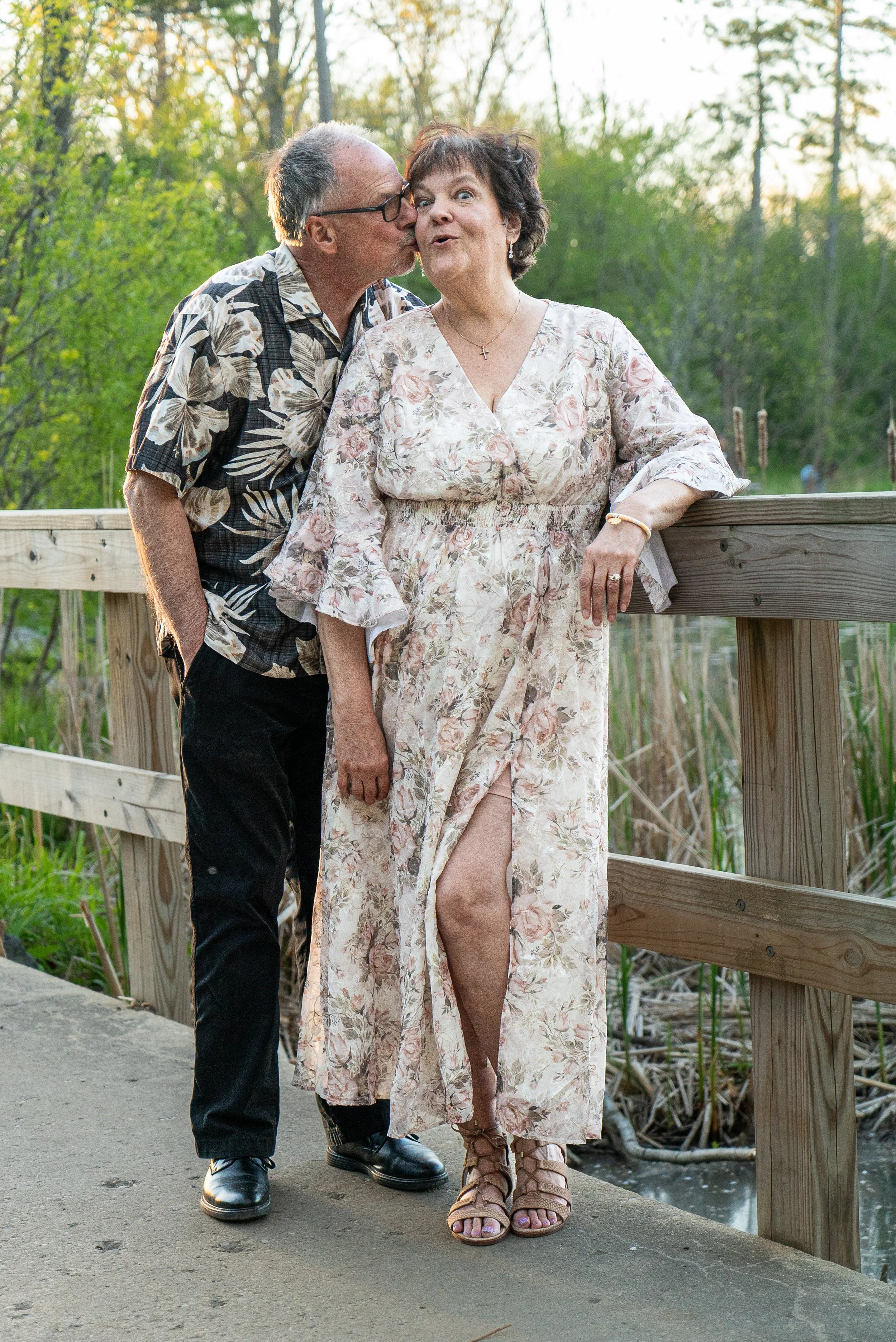 An elderly man kissing an elderly woman on the cheek on a wooden bridge outdoors during daytime. The woman is wearing a long, floral dress with a high slit, and the man is in a short-sleeve button-up shirt with a tropical print and black pants. Green