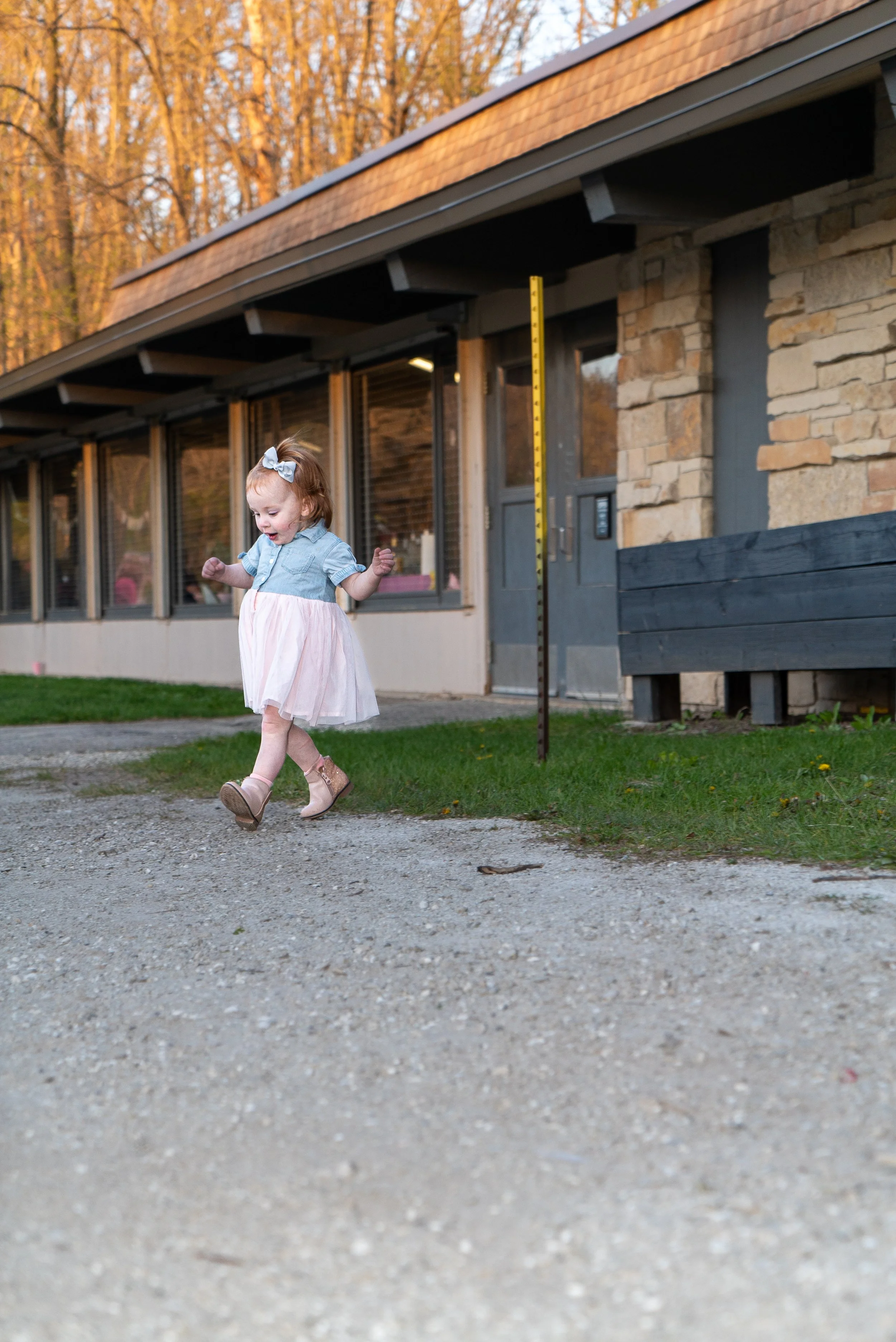 A young girl in a pink dress and denim jacket is joyfully running outside on a gravel path, with a building and trees in the background during sunset.