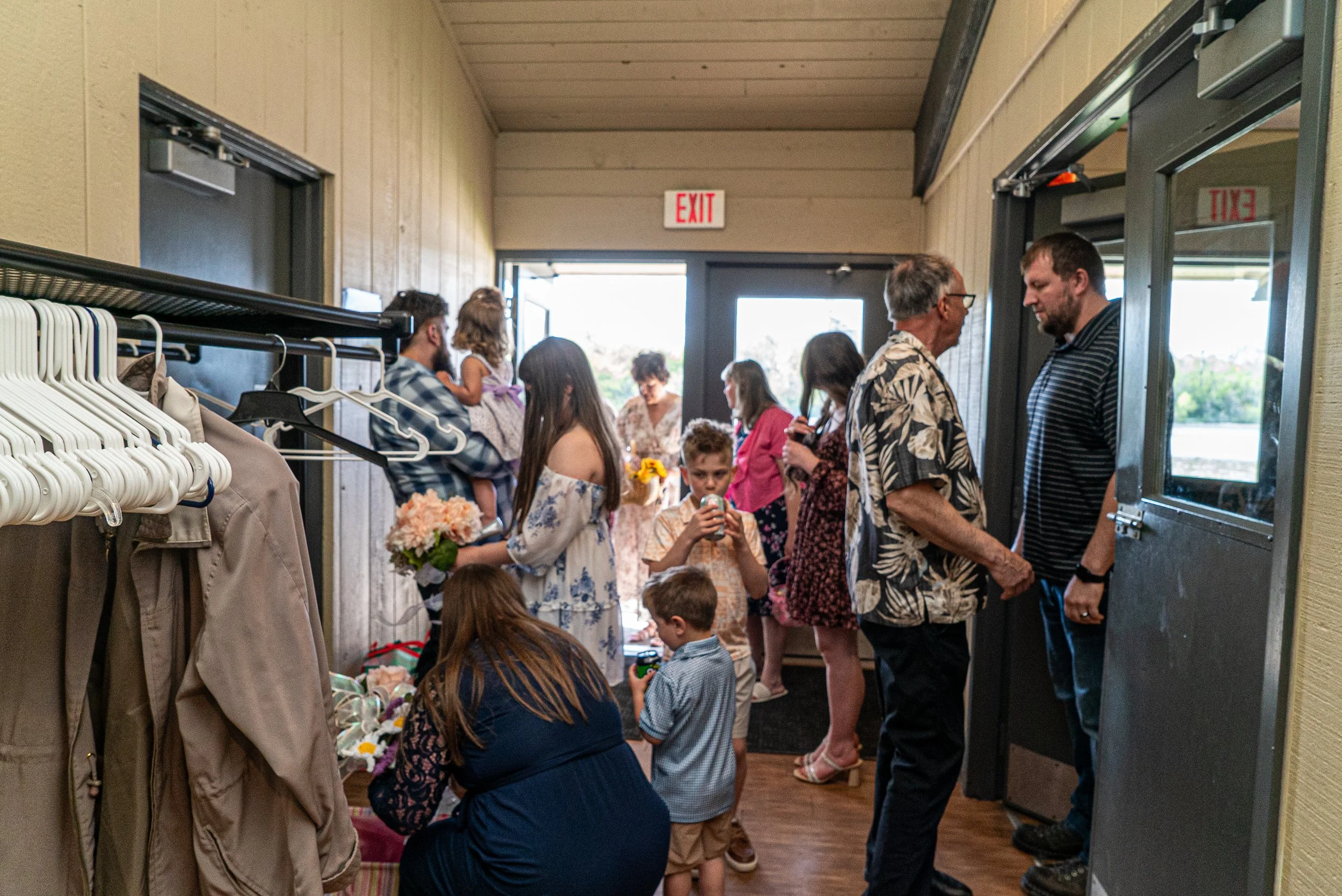 Group of people gathered indoors near an exit door, some sitting, some standing, engaged in conversation, with clothing racks on the left and a window showing an outdoor scene.