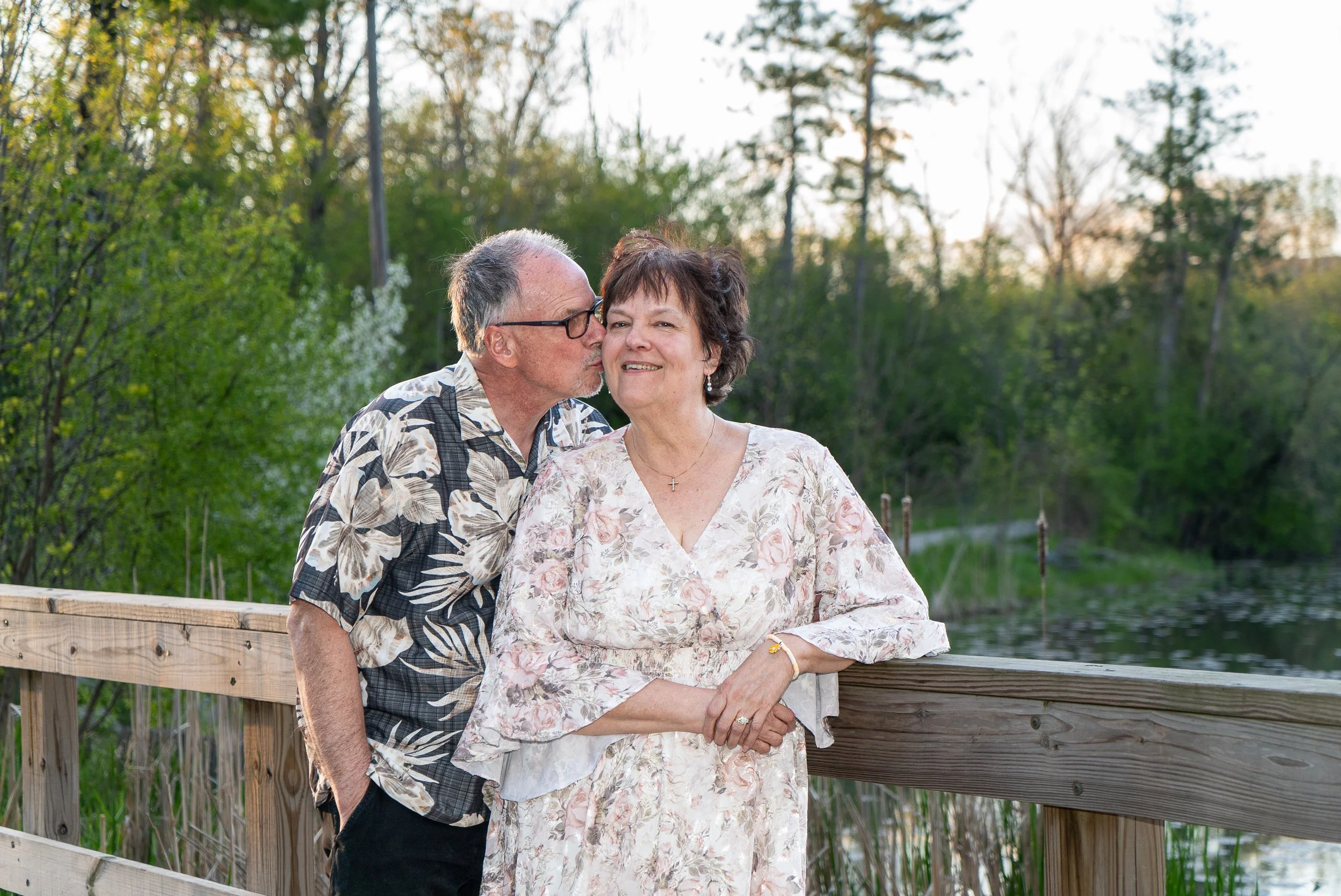 An elderly couple standing on a wooden bridge near a lake, with the man kissing the woman's cheek. The woman is smiling, wearing a floral dress, and the man is wearing glasses and a patterned shirt. There are trees in the background with green leaves
