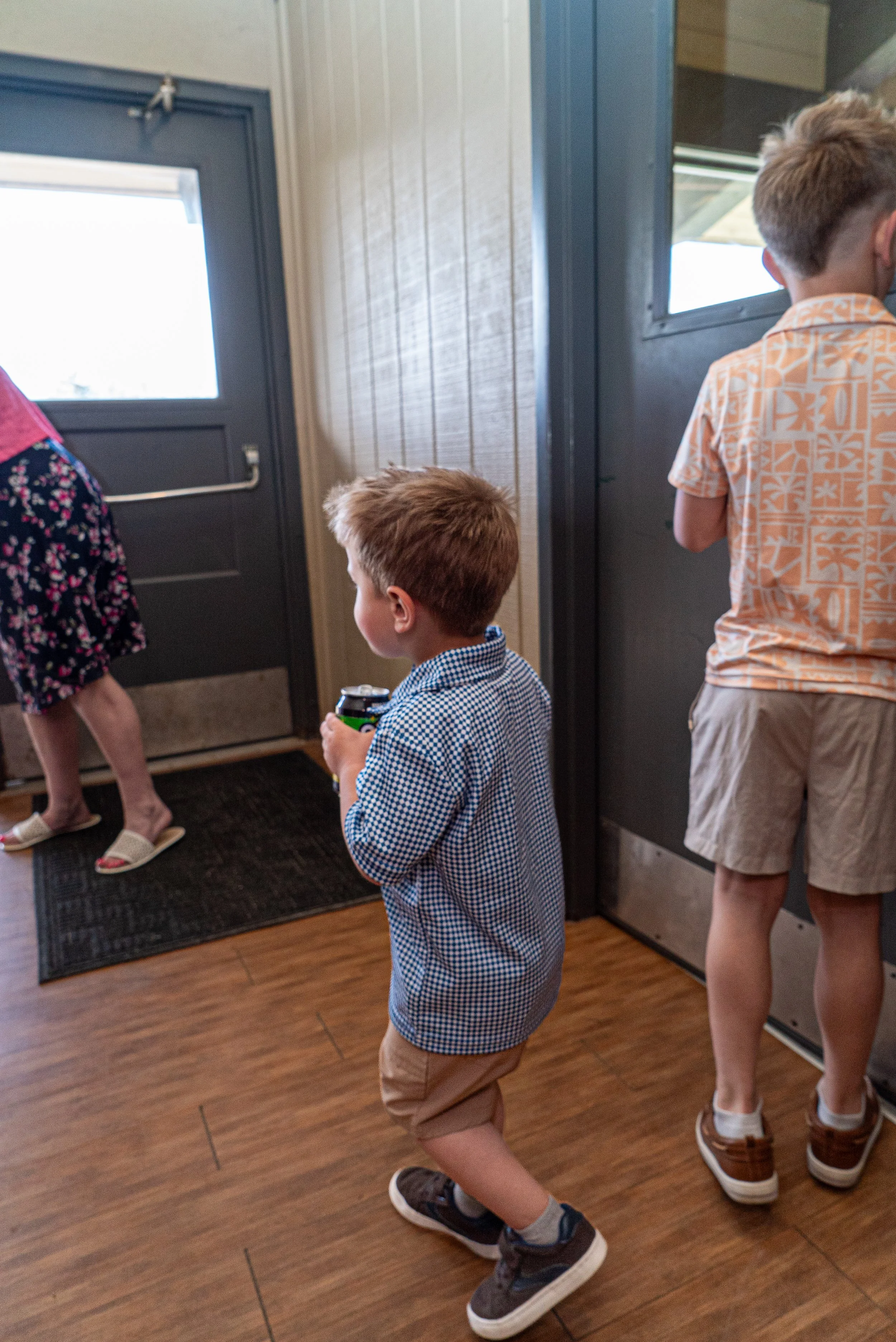 Children waiting indoors, some sitting or standing near a door with a window, wearing casual summer wedding clothing.