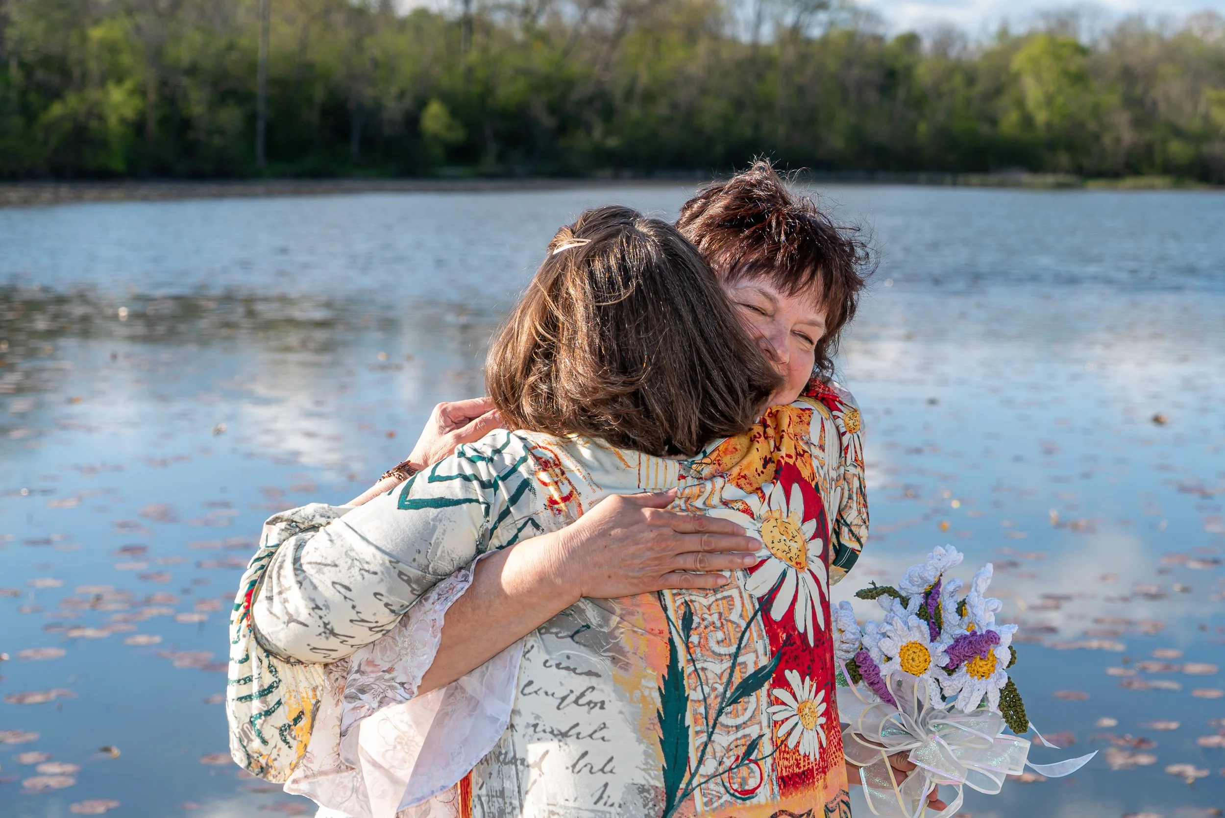 Two women embracing near a body of water with trees in the background, one holding a bouquet of flowers.