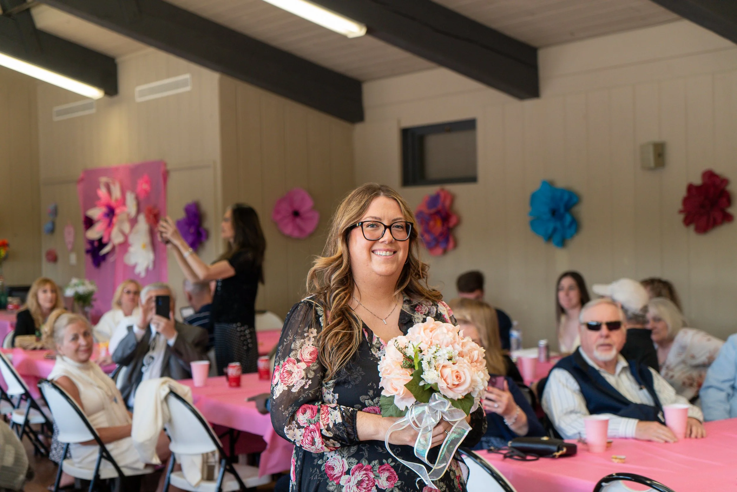 A woman with glasses and long wavy hair holding a bouquet of pink and white flowers at a celebration in a decorated hall with pink and purple wall decorations, and many seated guests.