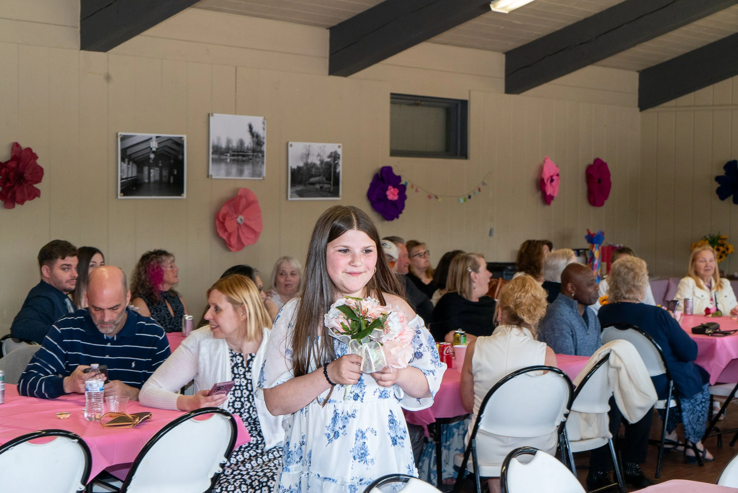 A young girl holding a bouquet of flowers standing in front of a group of seated people at a table with pink tablecloths during a gathering or celebration in a decorated hall.