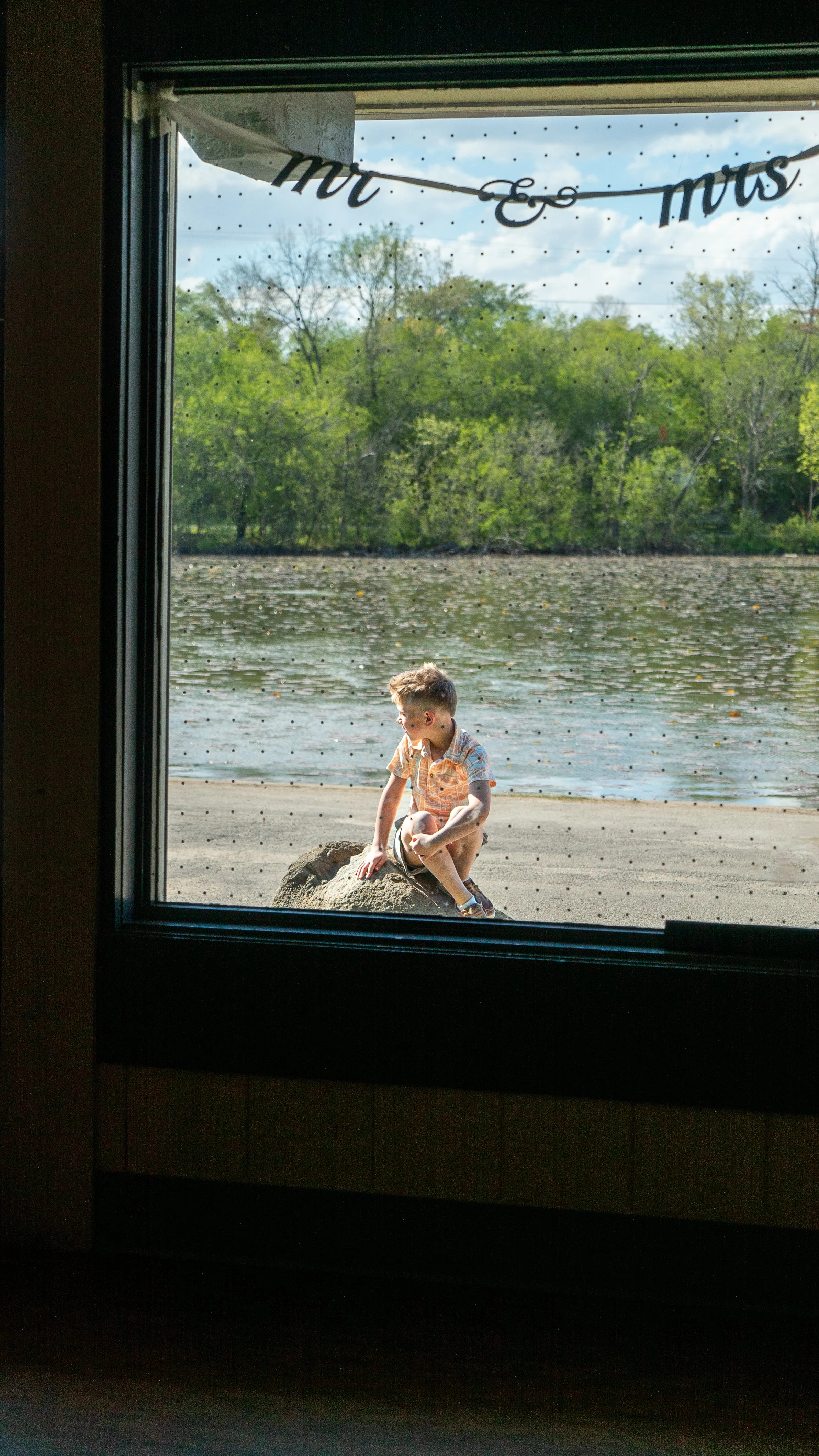 View through a window showing a young boy sitting on a rock near a river, with trees and a blue sky in the background, and a decorative banner hanging inside.