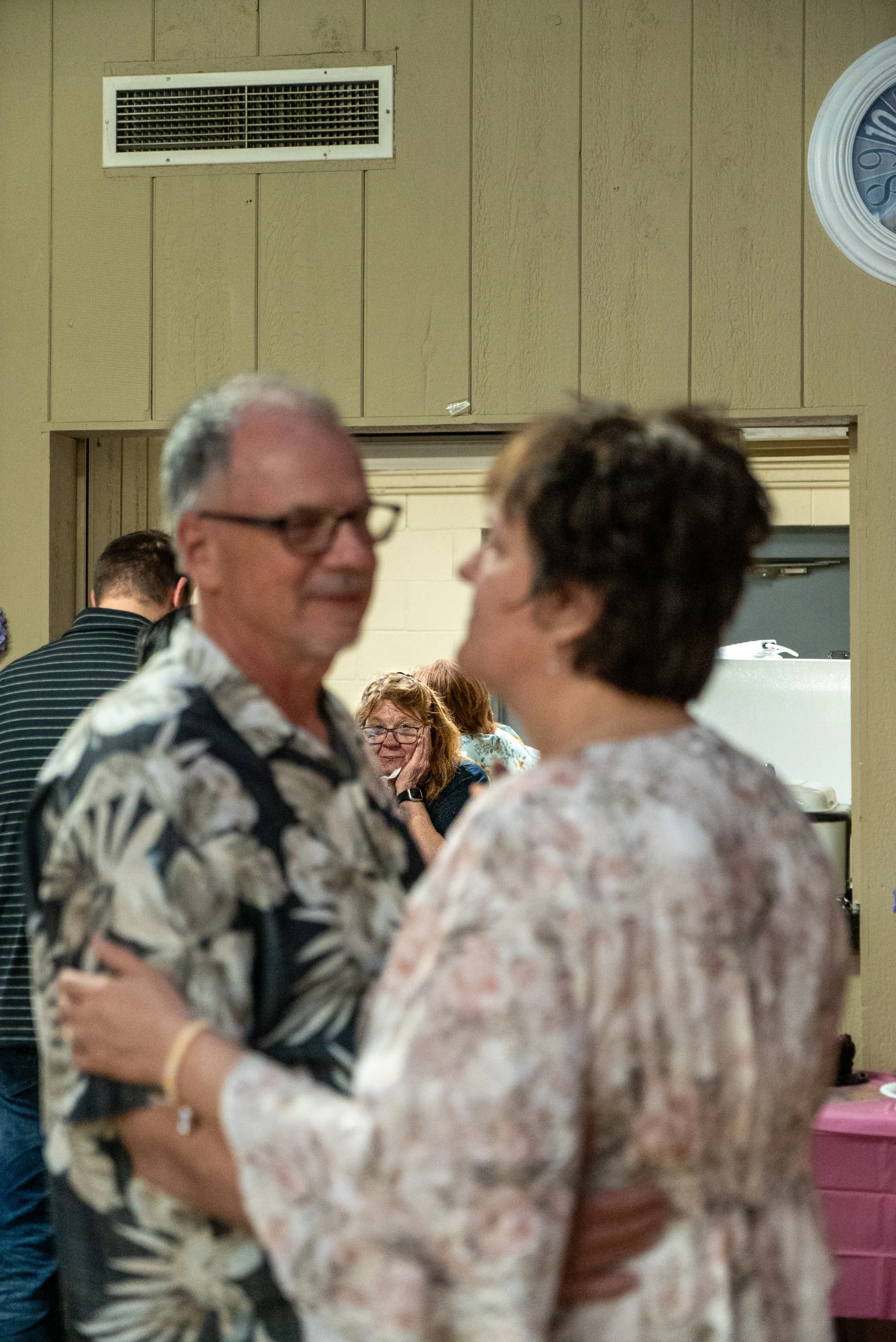Two women are standing close and holding hands, smiling at each other during an indoor event. People are visible in the background, engaged in conversations.