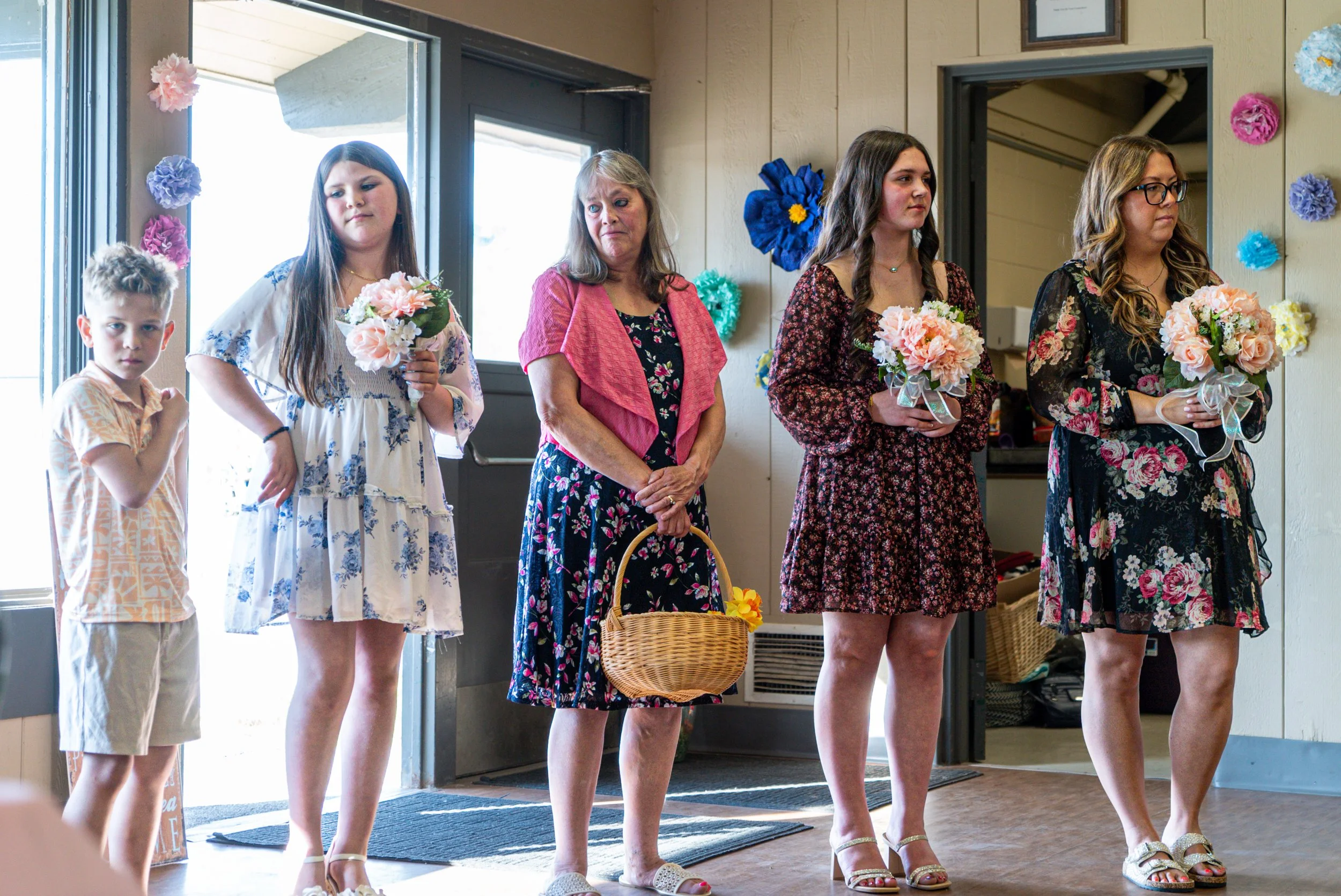 A group of five women and a young boy standing in a line indoors, holding bouquets of flowers, with colorful paper flowers decorating the walls behind them.