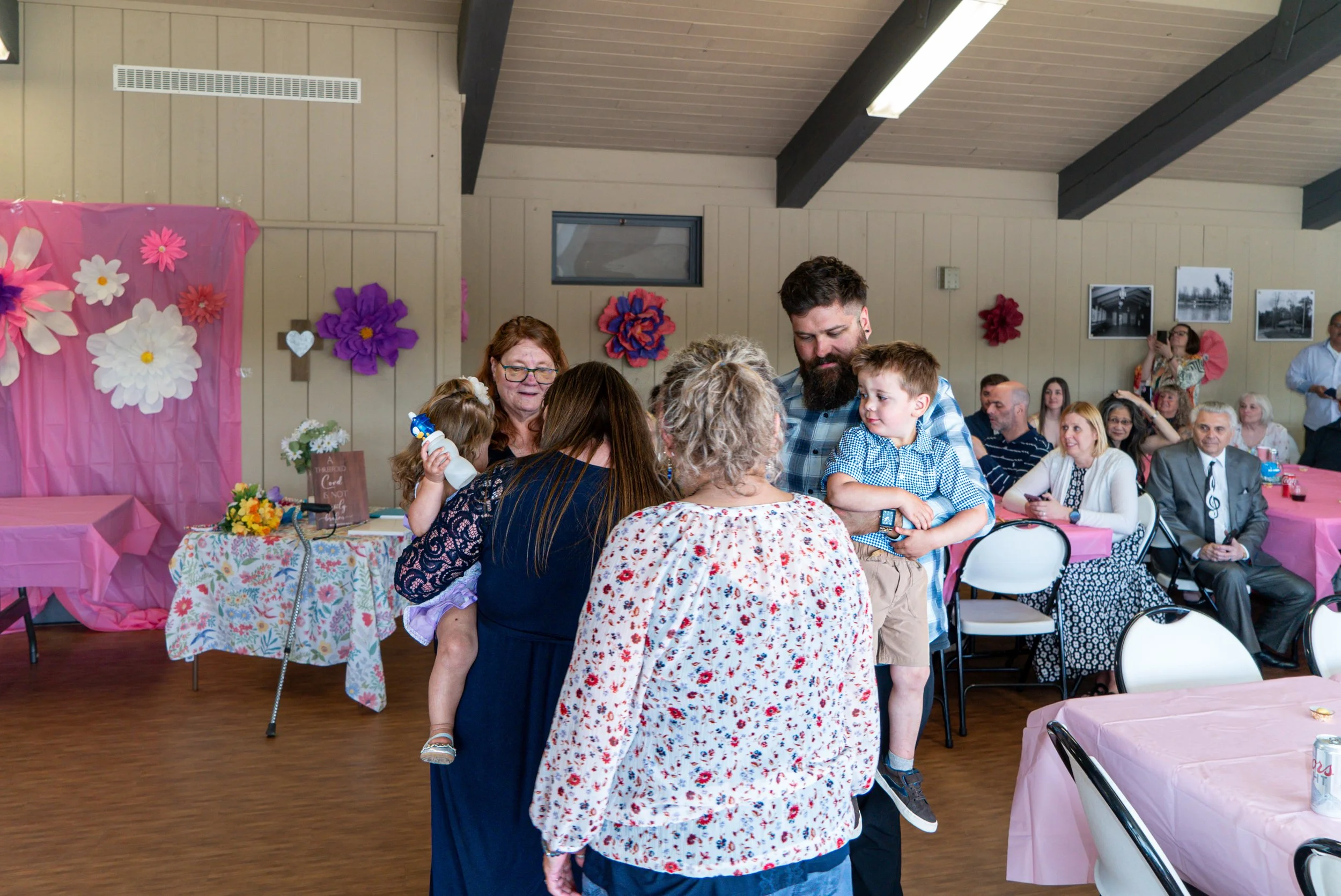 People gather around a table with pink tablecloths and floral decorations at a celebration in a decorated hall, with others seated at tables in the background.