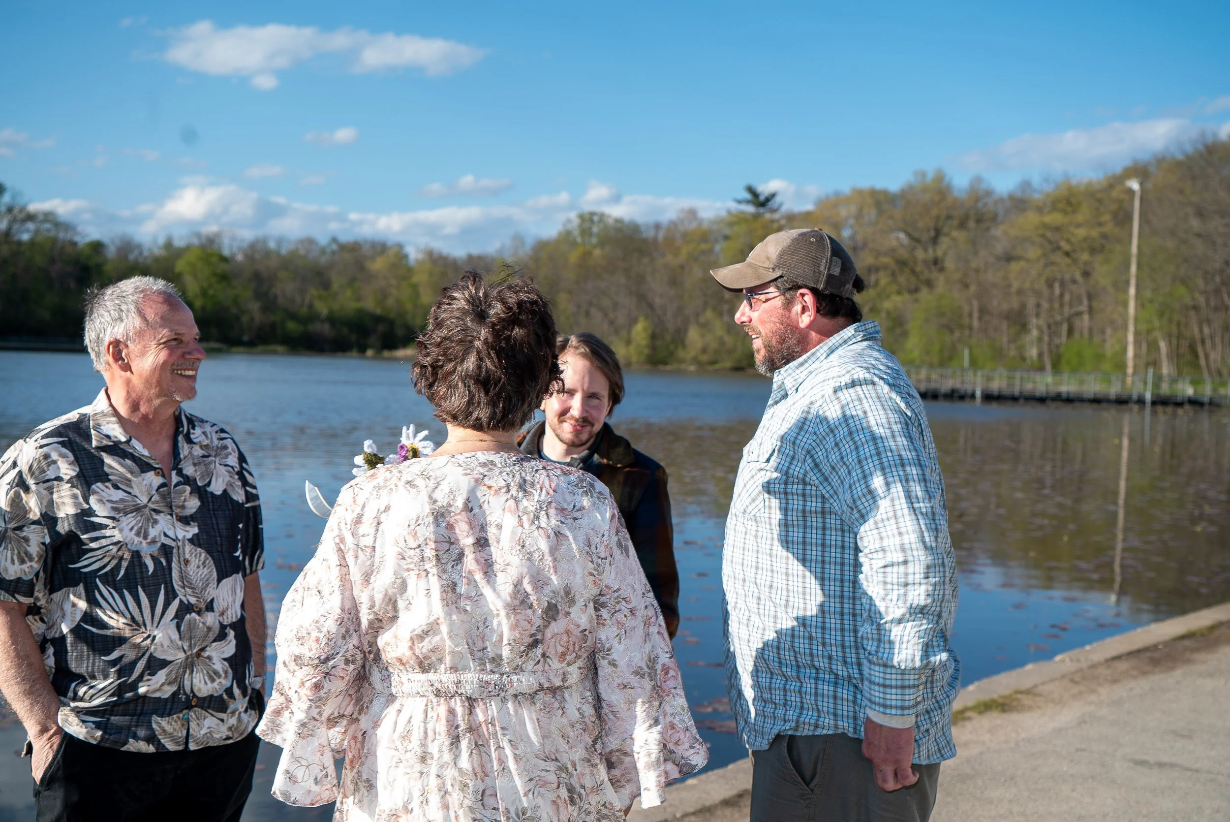 A group of five people standing outdoors near a body of water, engaged in conversation on a sunny day. The group includes three men and two women, with trees and a clear blue sky in the background.