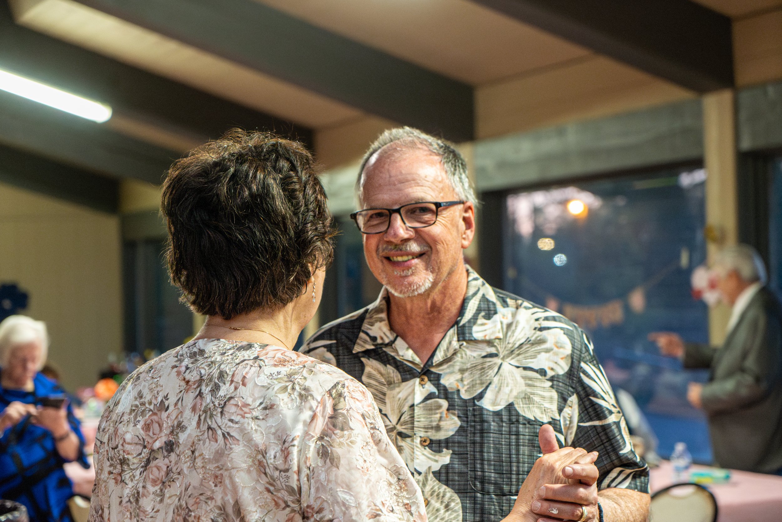 A man and a woman dancing closely together at an indoor social event, both wearing patterned Hawaiian shirts.