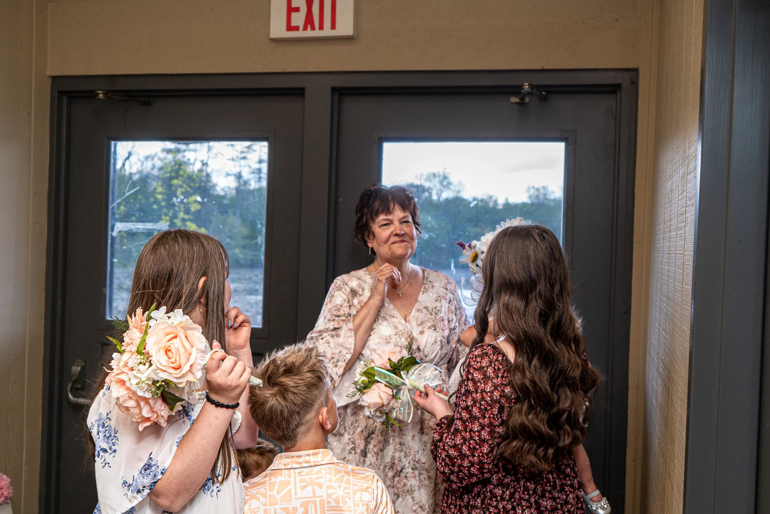 A group of young girls and an older woman, possibly a grandmother, in front of a door with an 'EXIT' sign above it, holding flower bouquets and engaging in conversation or a celebration.