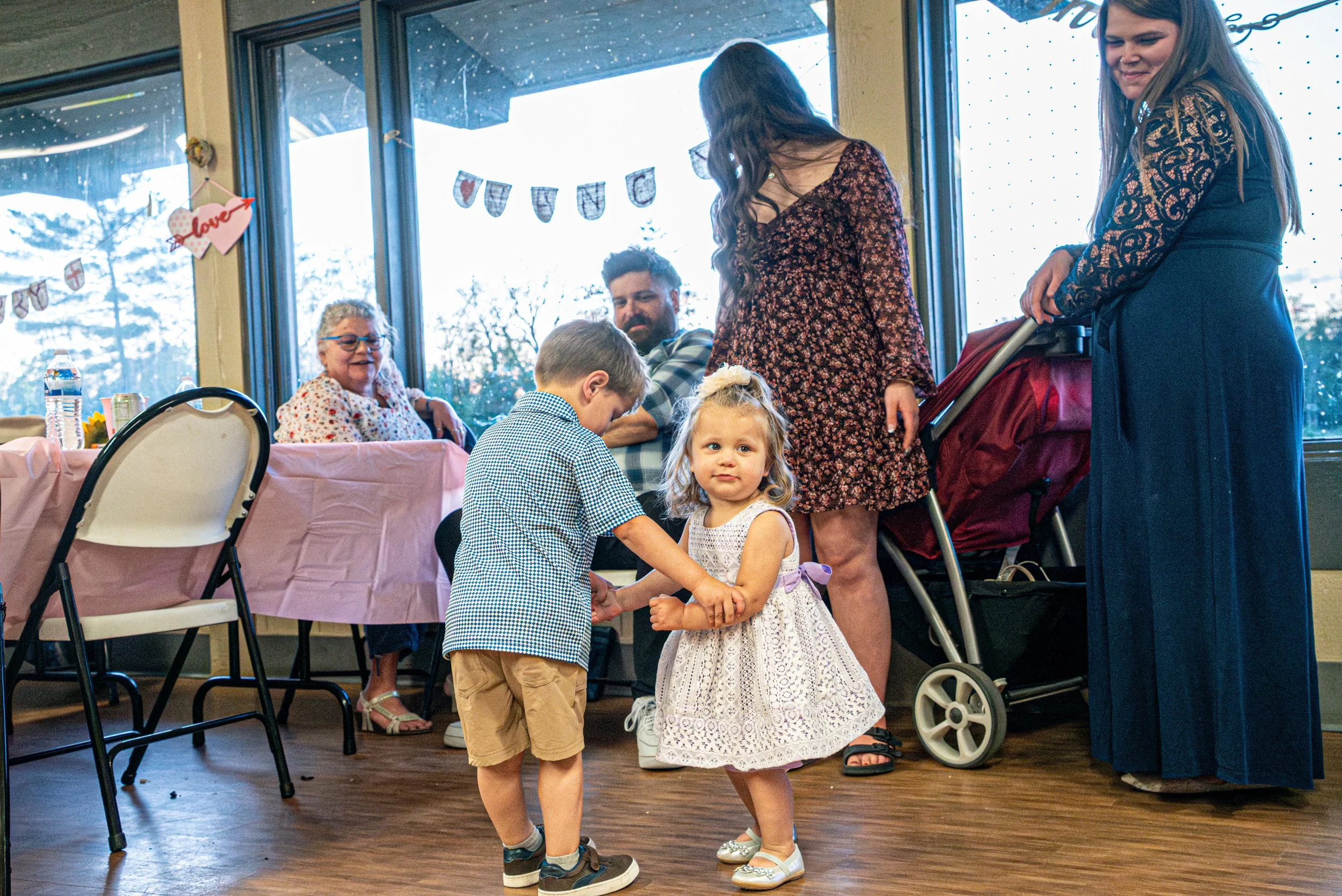Two young children holding hands and dancing in front of a group of adults at a celebration, with a decorated and well-lit room in the background.