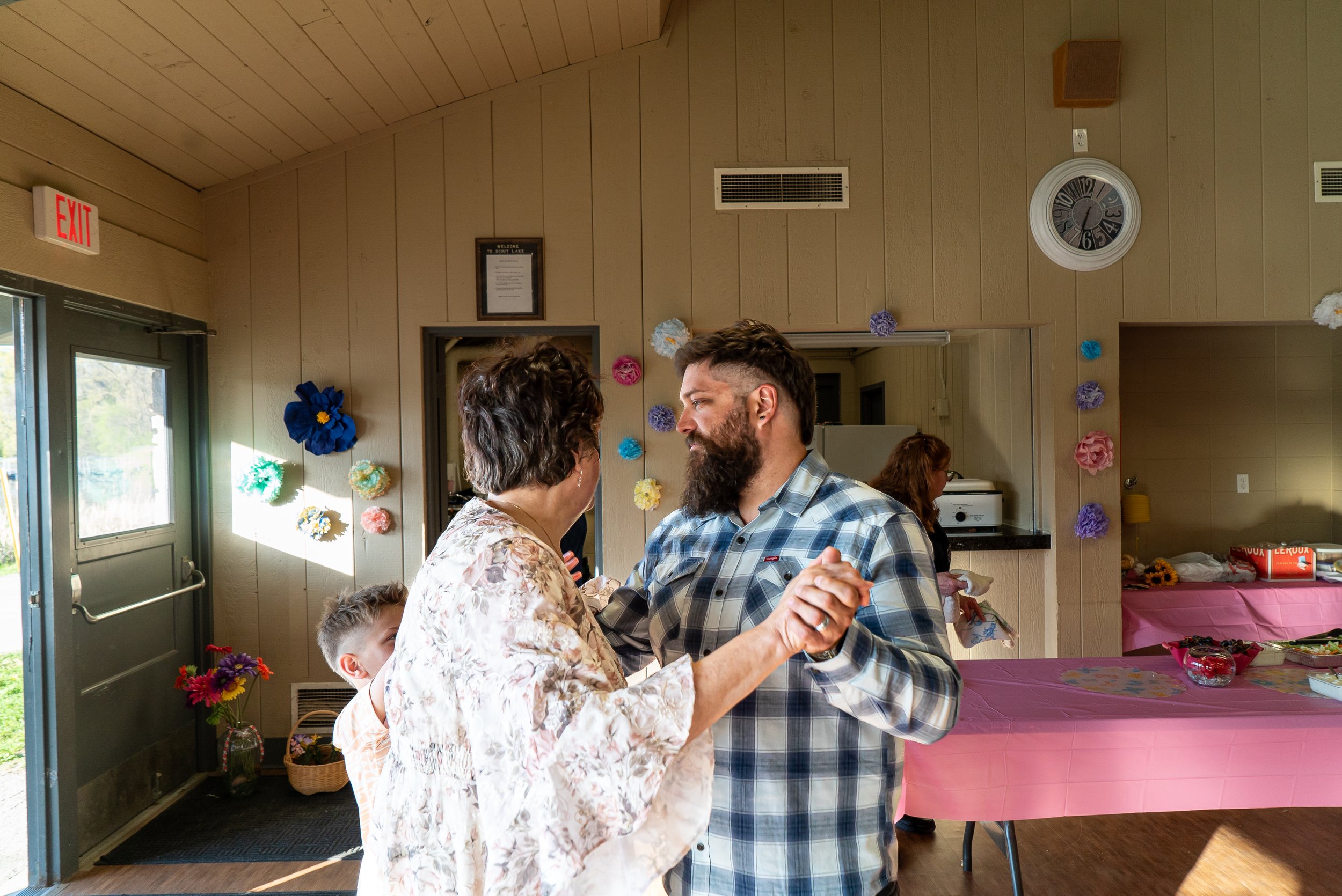 A couple dancing together at a celebration in a decorated hall with floral decorations on the wall and a table with a pink tablecloth.