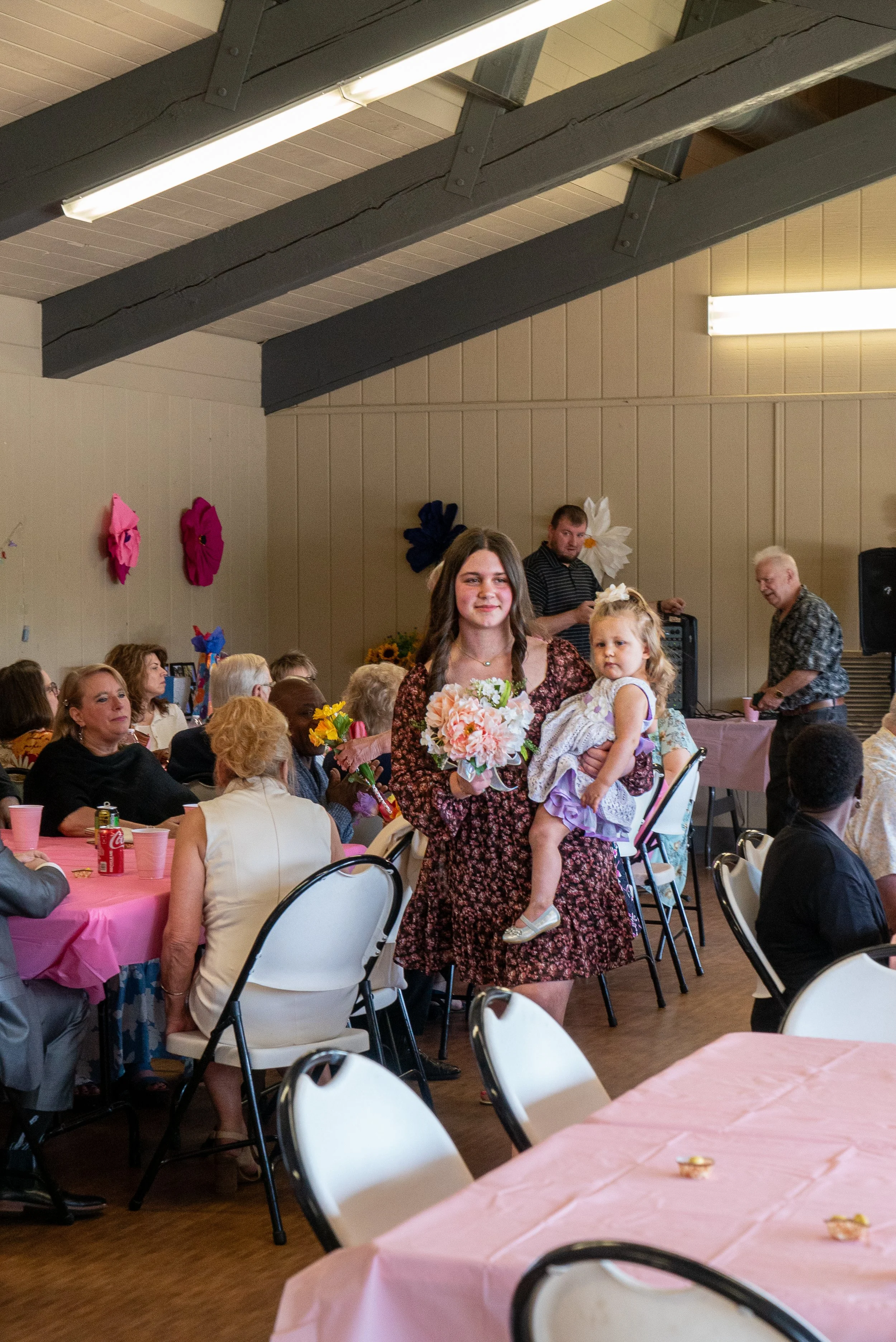 A woman holding a young girl in a gathering or party setting with many people seated at tables covered with pink tablecloths, and ornaments on the walls.
