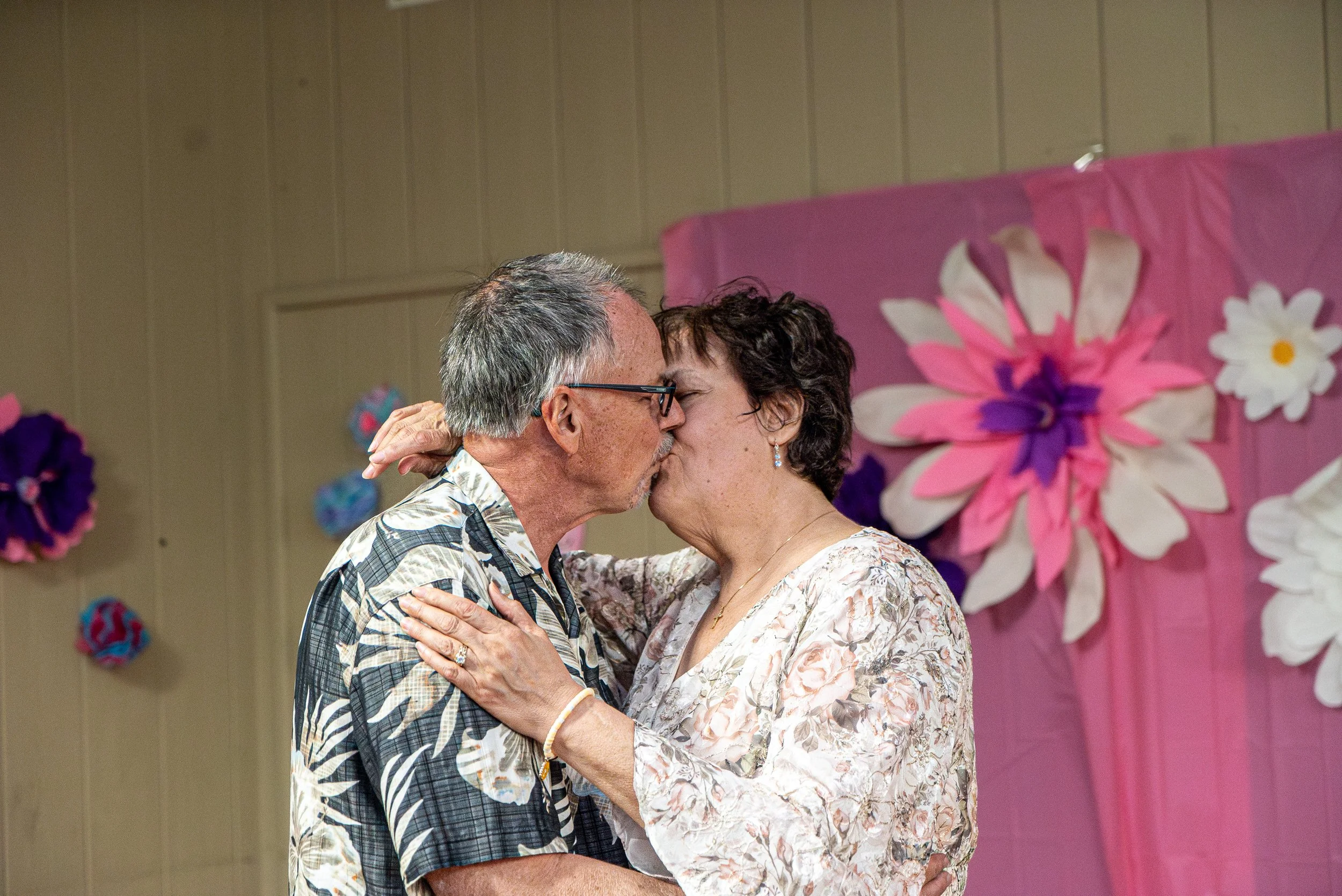 An elderly couple dancing closely, about to kiss, in front of colorful paper flower decorations on a wall.