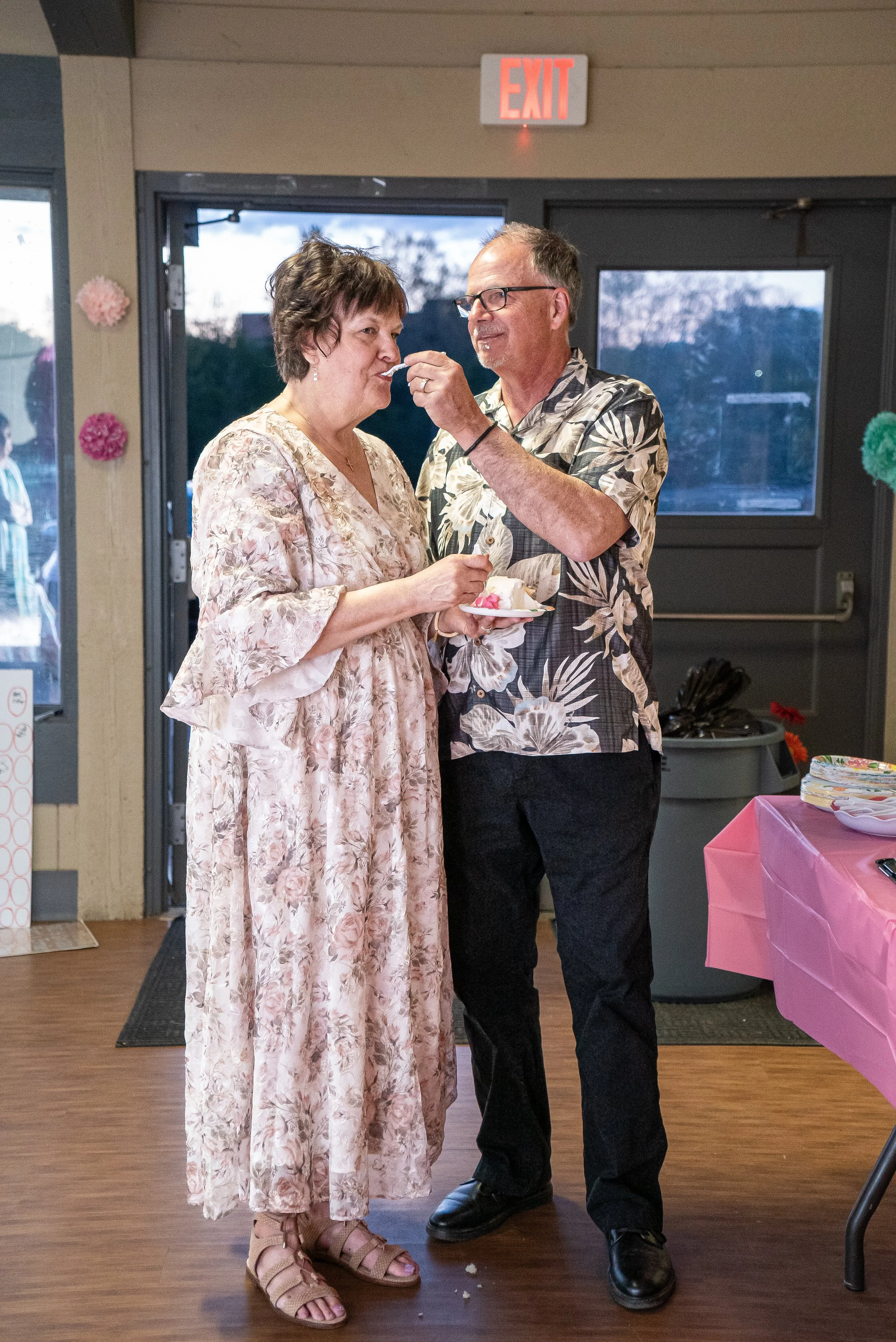A man helping a woman eat cake at a celebration or party indoors, near an exit sign and decorated with pink paper flowers.
