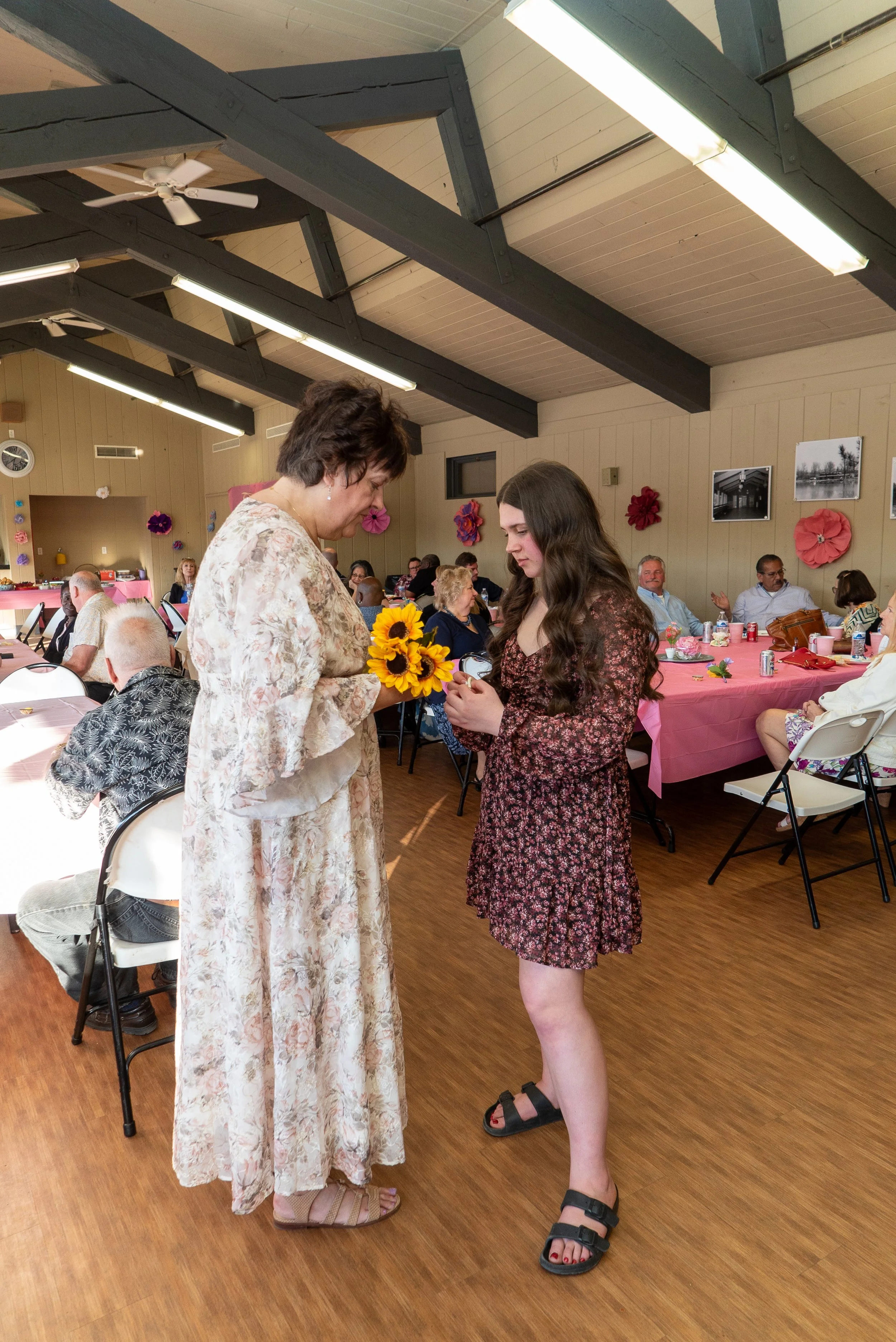A woman in a floral dress and another woman in a floral dress exchanging rings during a ceremony in a decorated hall with pink tablecloths, flower decorations, and seated guests.