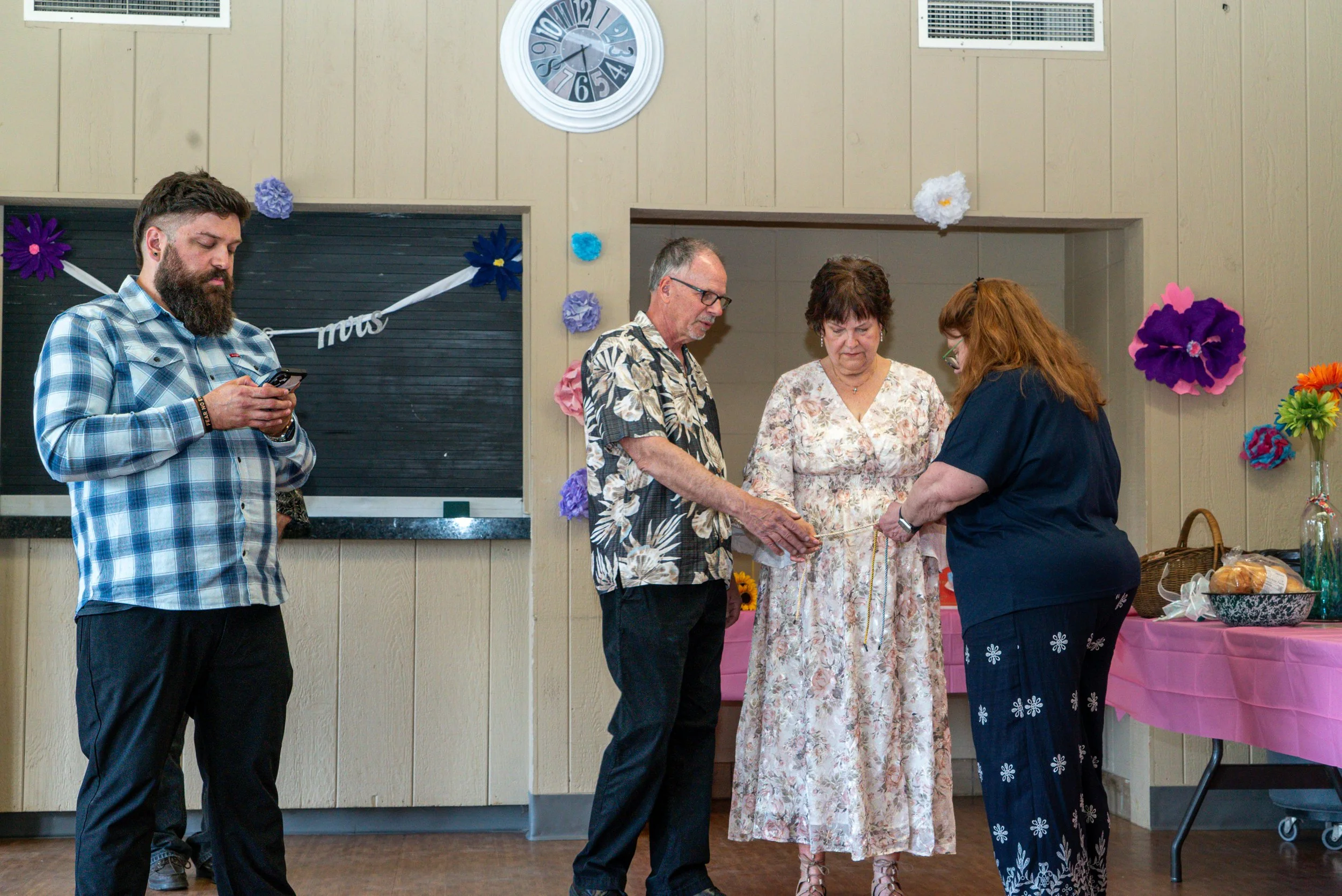 A group of five people standing indoors, with three engaged in a handshake and conversation. One person on the left is looking at their phone. The background has beige wood-paneled walls, colorful paper flowers, a round clock, and a table with a pink