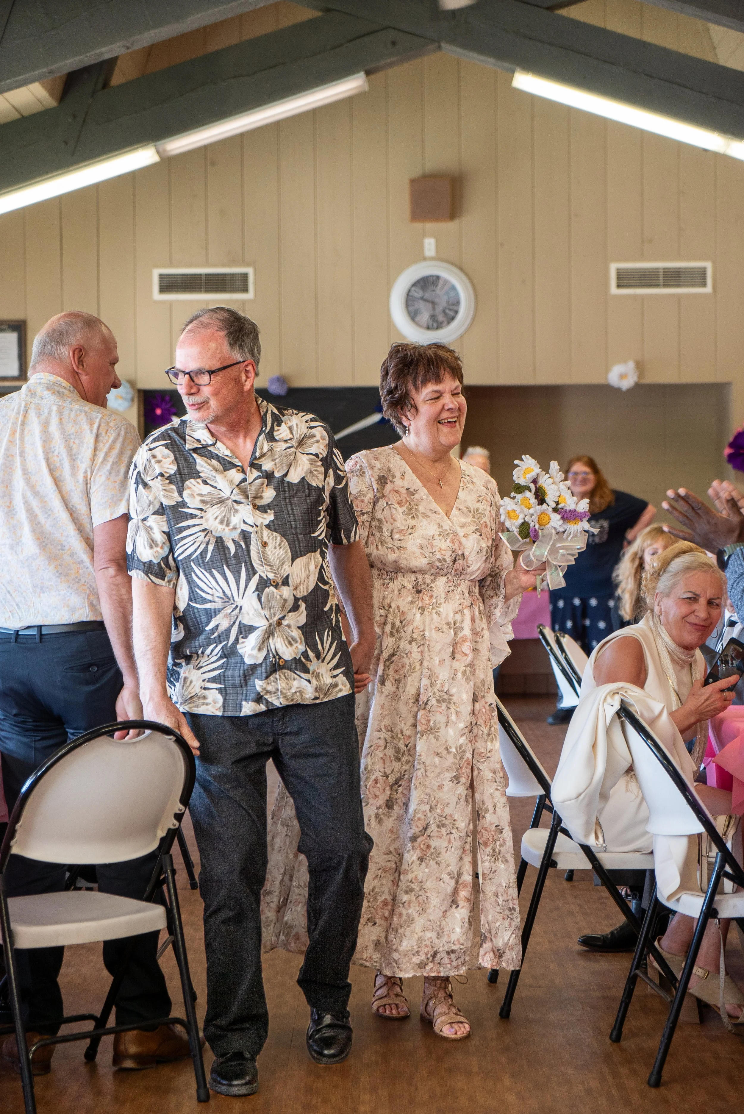 People gathered in a room, some standing and some seated, in a celebration or social event. A woman in a floral dress holds a bouquet of flowers and is smiling, with other people around her.