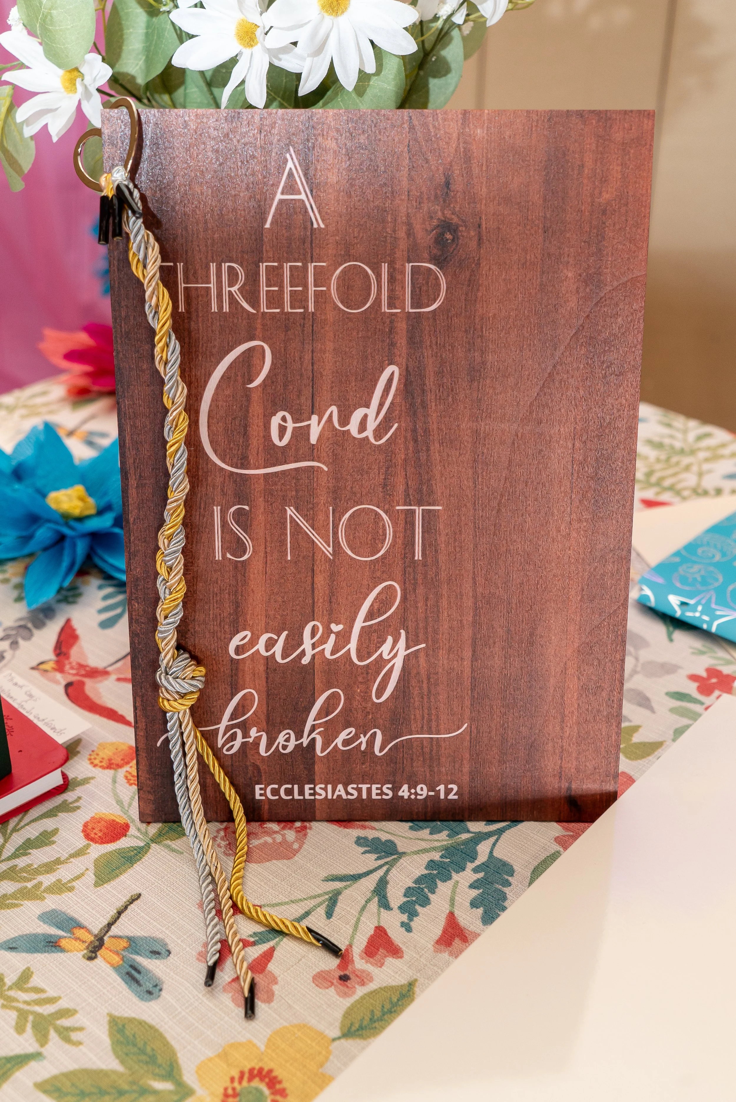 A wooden sign with an inspirational quote from Ecclesiastes 4:9-12, decorated with a twisted gold, silver, grey, and black cord, standing on a table with a floral tablecloth and a bouquet of white daisies in the background.