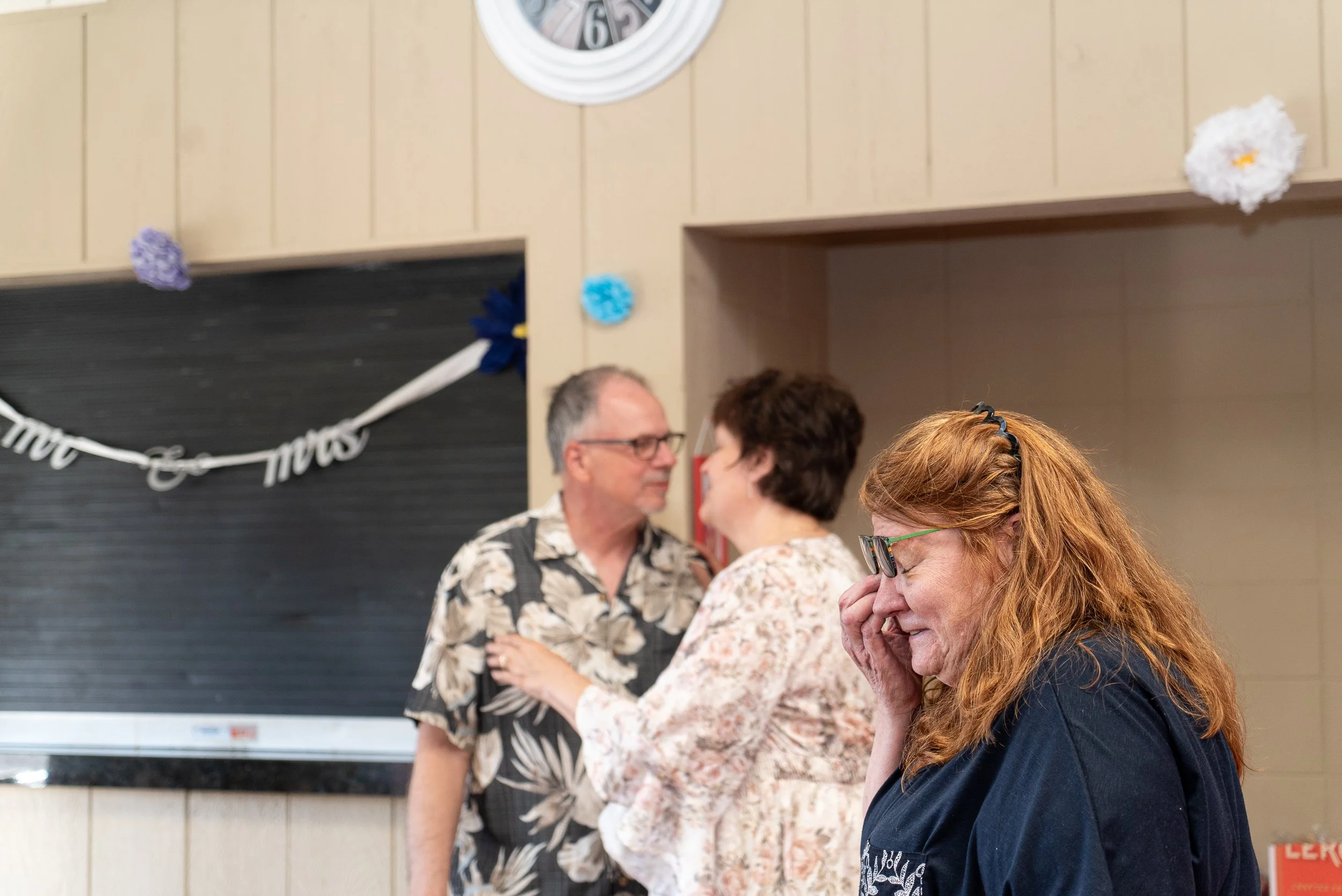 Two women and two men smiling and talking to each other indoors, with a decorative clock and paper flower decorations on the wall in the background.
