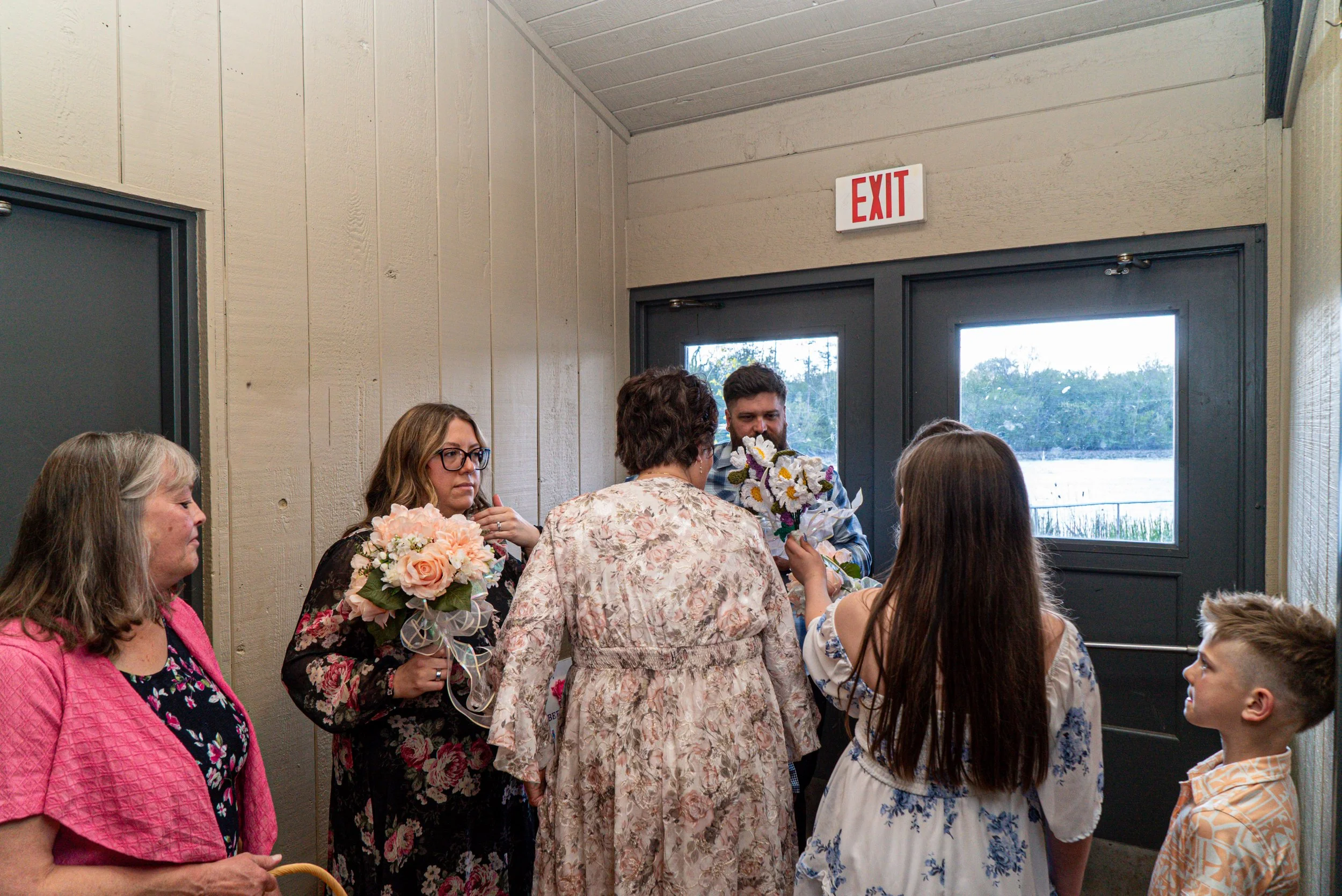 Group of people, mostly women, indoors near exit door, some holding bouquets of flowers, engaged in conversation.