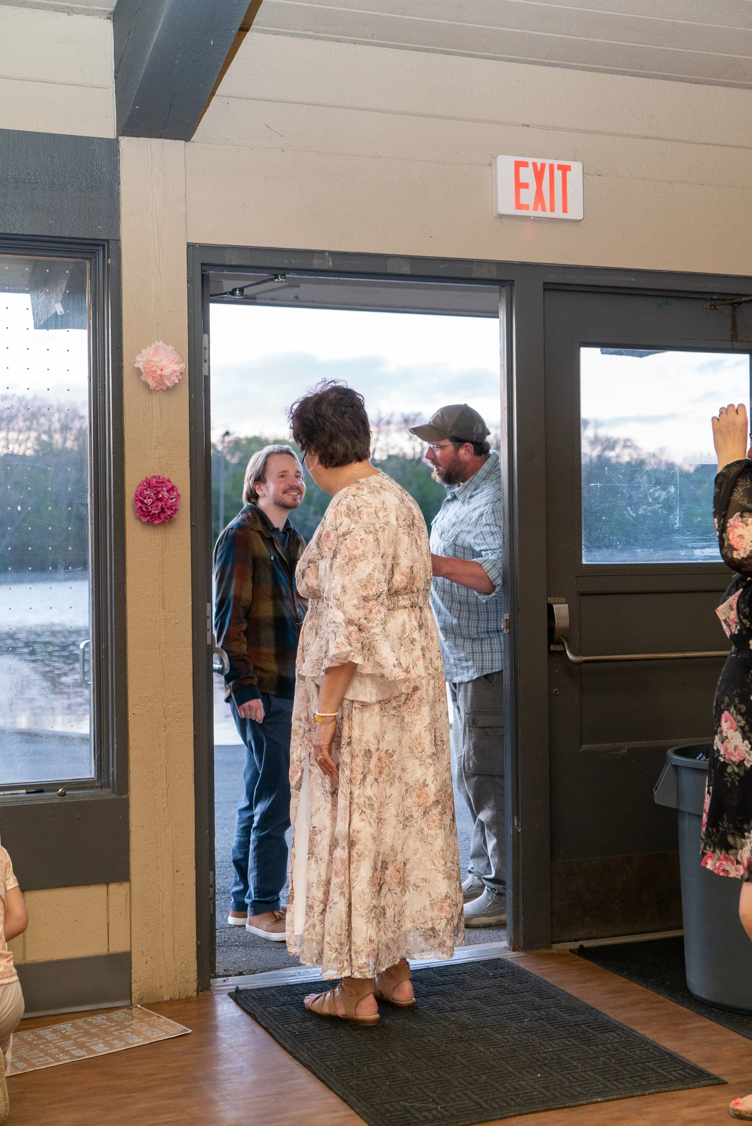 People gathered at the entrance of a building, engaging in conversation. A woman in a floral dress and sandals is standing at the door, with others nearby. The scene is indoors near the doorway, with decorative paper flowers on the wall and a view of