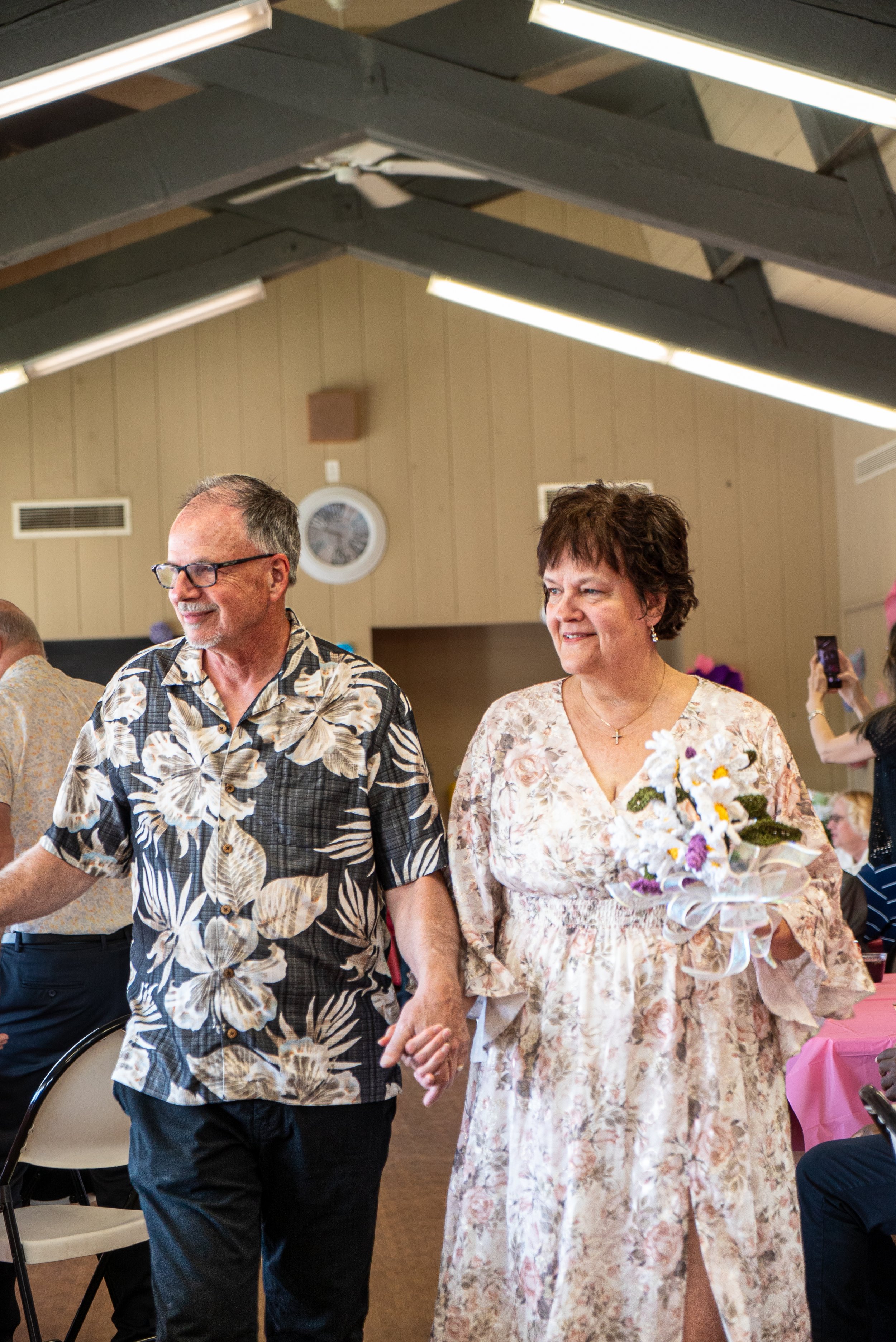 A couple holding hands, walking in a gathering, with the woman holding a bouquet of flowers, in an indoor space decorated for a celebration.