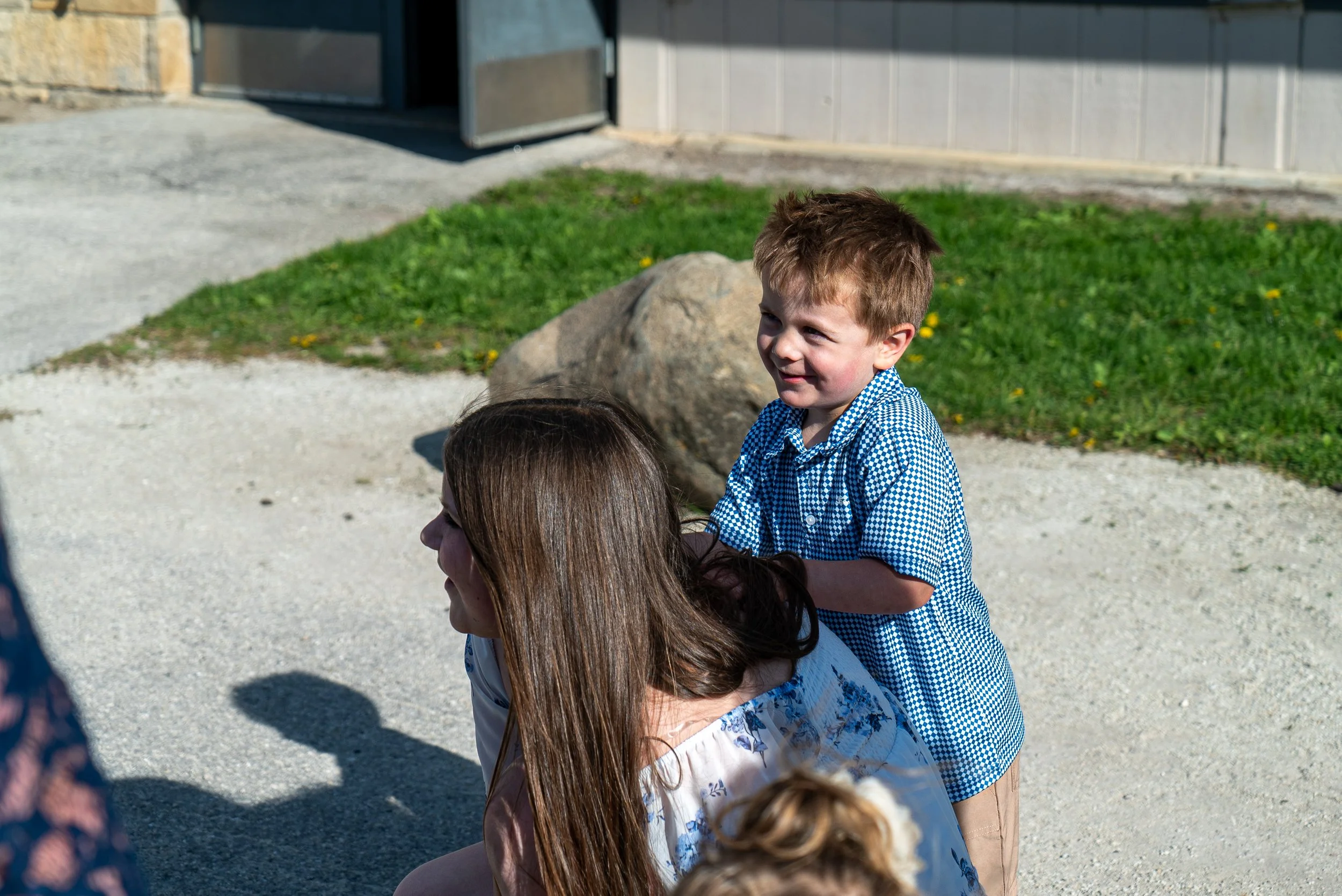 A young boy with light brown hair, wearing a blue and white checkered shirt, sitting on a woman's shoulders outdoors. The woman, with long brown hair, is partially visible. They are near a grassy area with a rock and a building in the background.