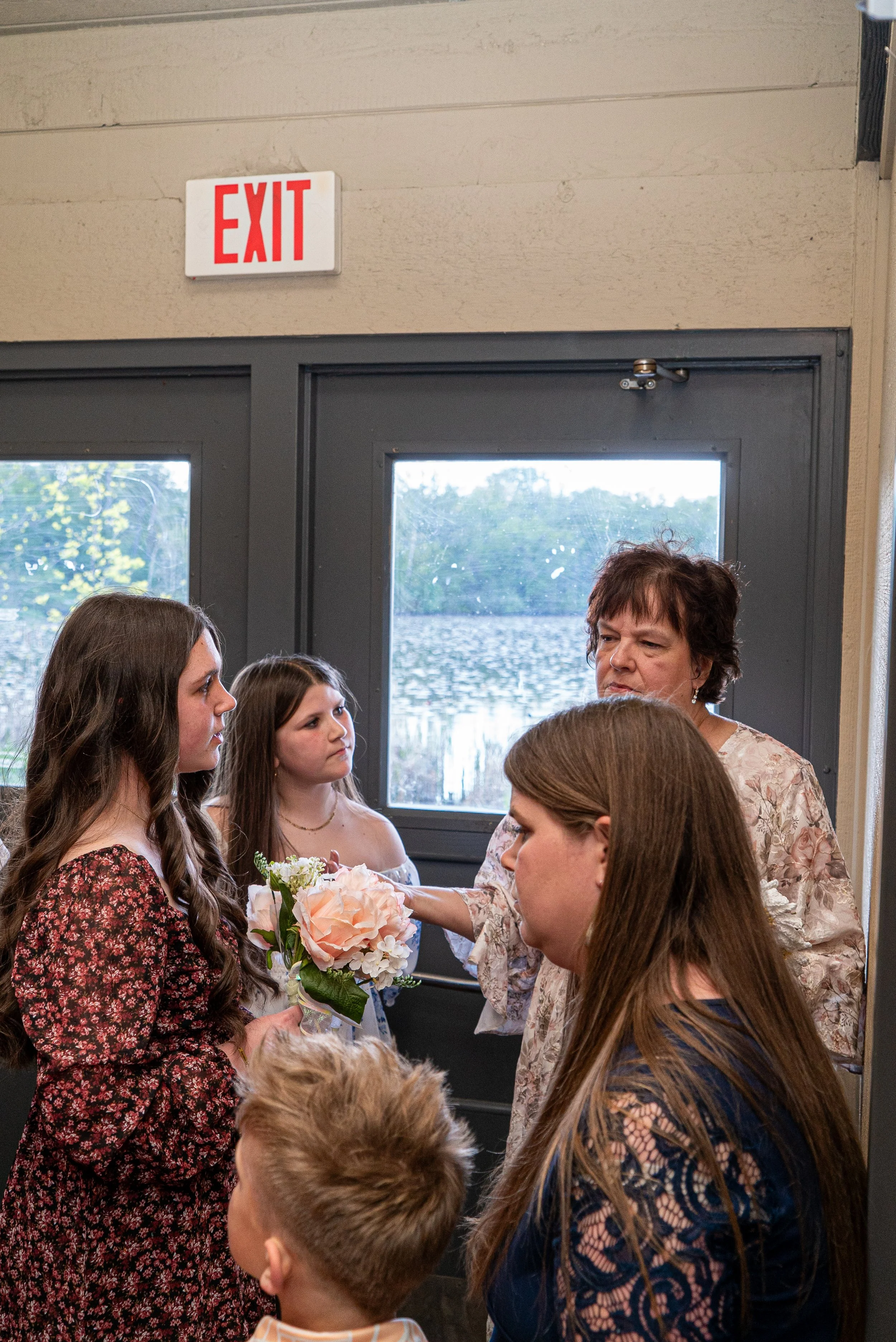 People gathered indoors near a wall with a window, with an exit sign above the door. A woman is handing a bouquet of flowers to a young woman with long, curly hair.